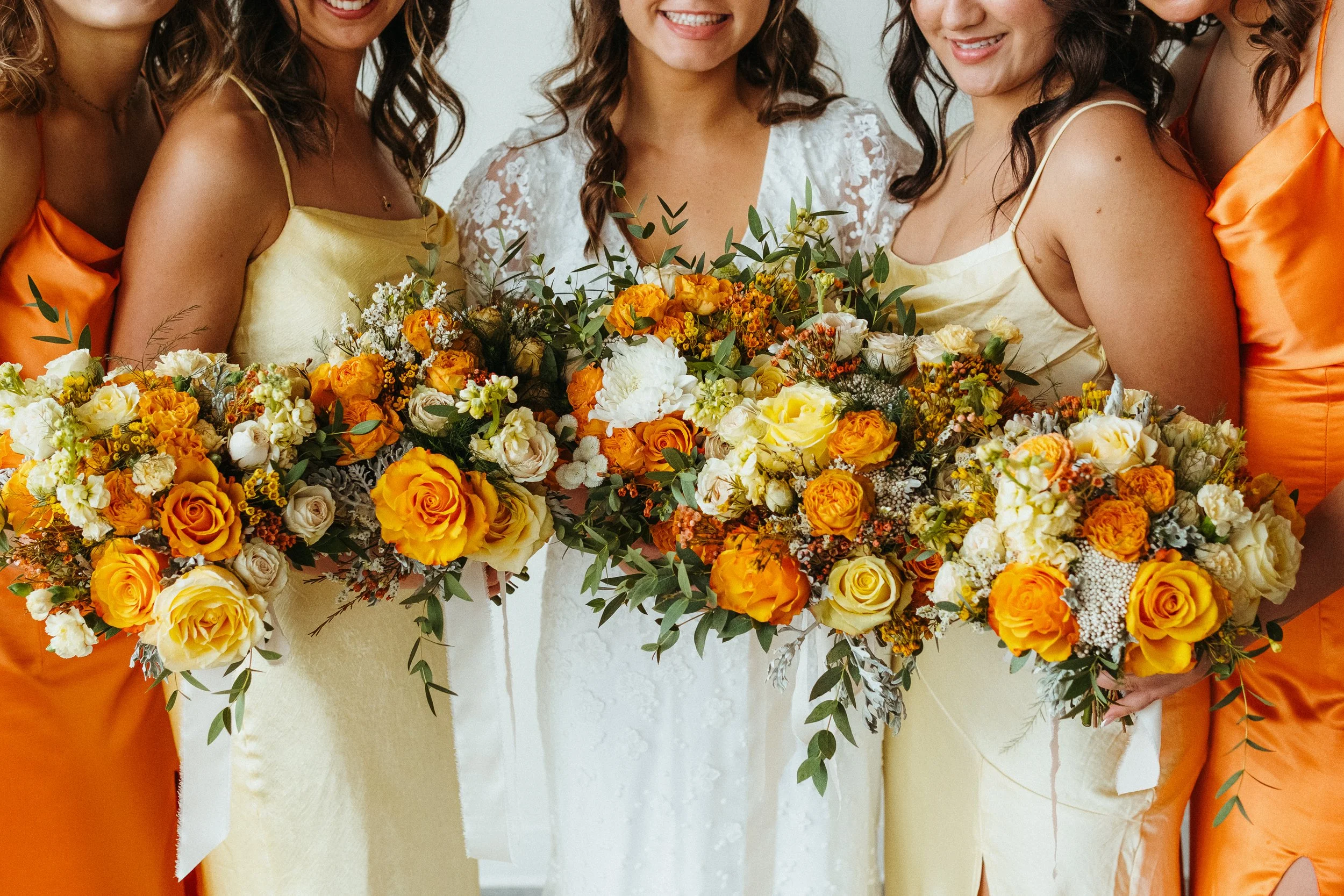 A group of women at a wedding holding large bouquets of yellow and orange flowers, with some wearing yellow dresses and others wearing orange dresses.