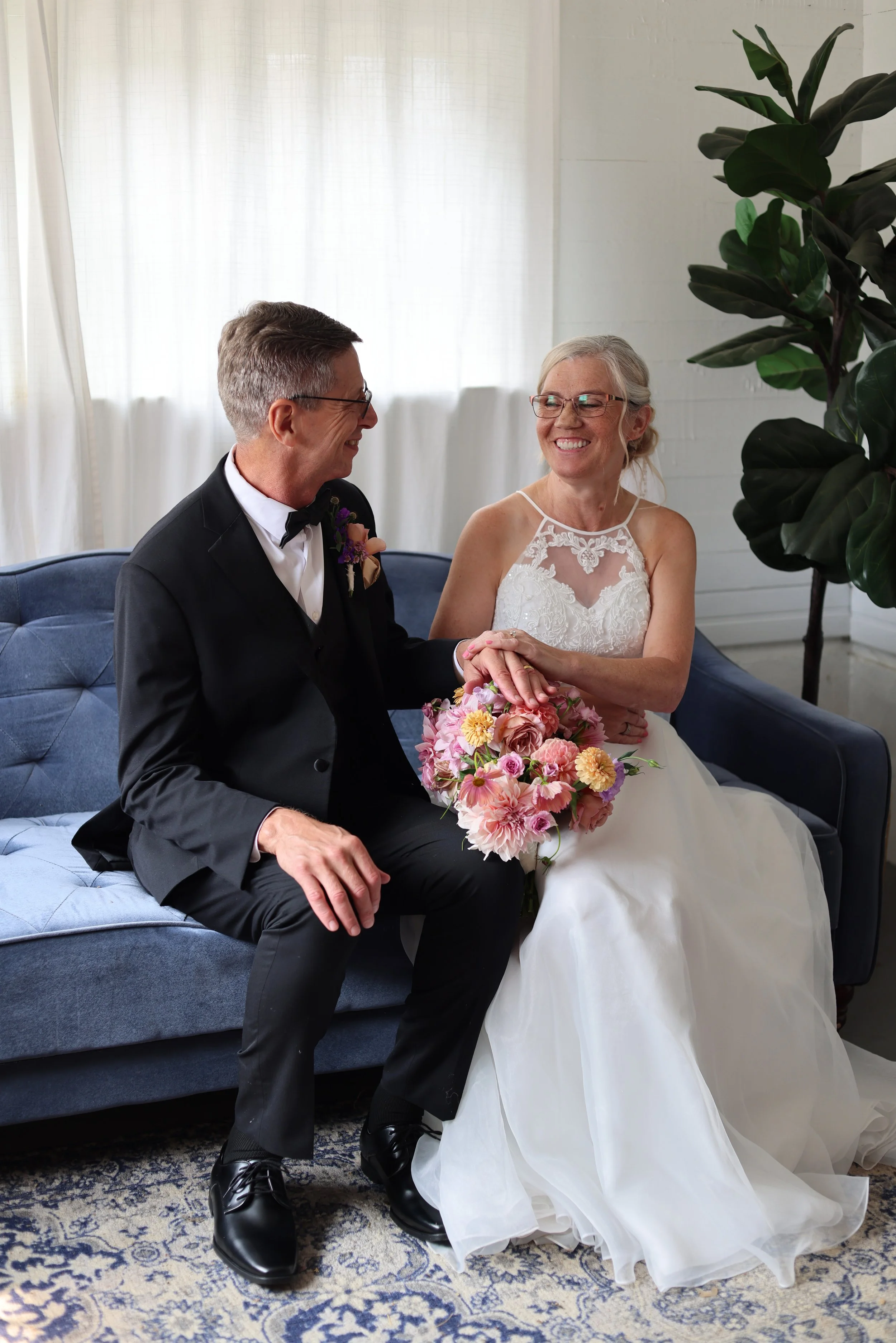 A bride and groom sitting on a blue sofa, holding hands and smiling at each other. The bride is in a white wedding dress holding a pink and yellow bouquet, and the groom is in a black tuxedo with glasses. There is a large green potted plant behind th