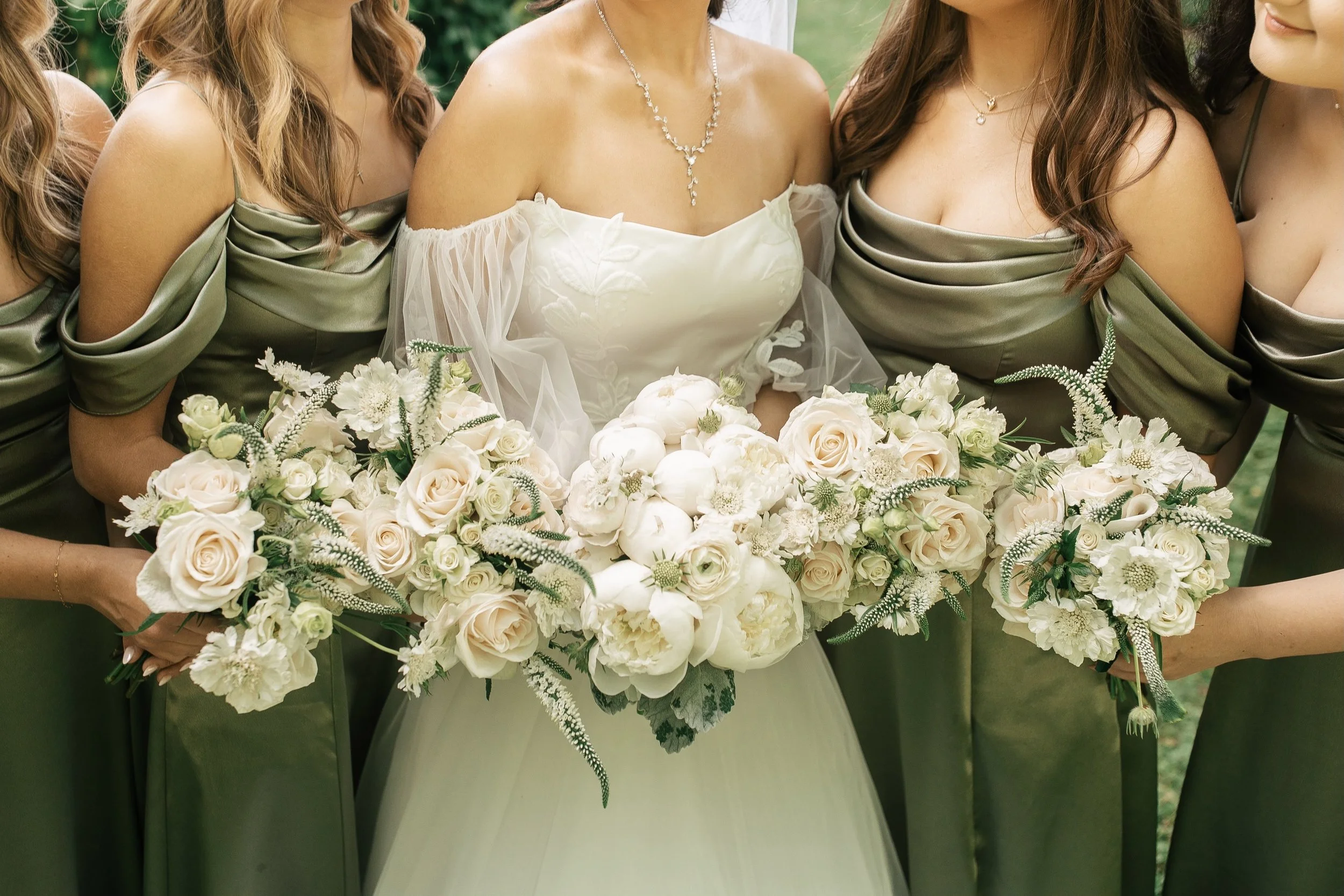 A bride in a white wedding gown holding a large bouquet of white and cream roses and flowers, standing among four bridesmaids in green satin dresses, each holding a similar smaller bouquet. The photo is taken outdoors.