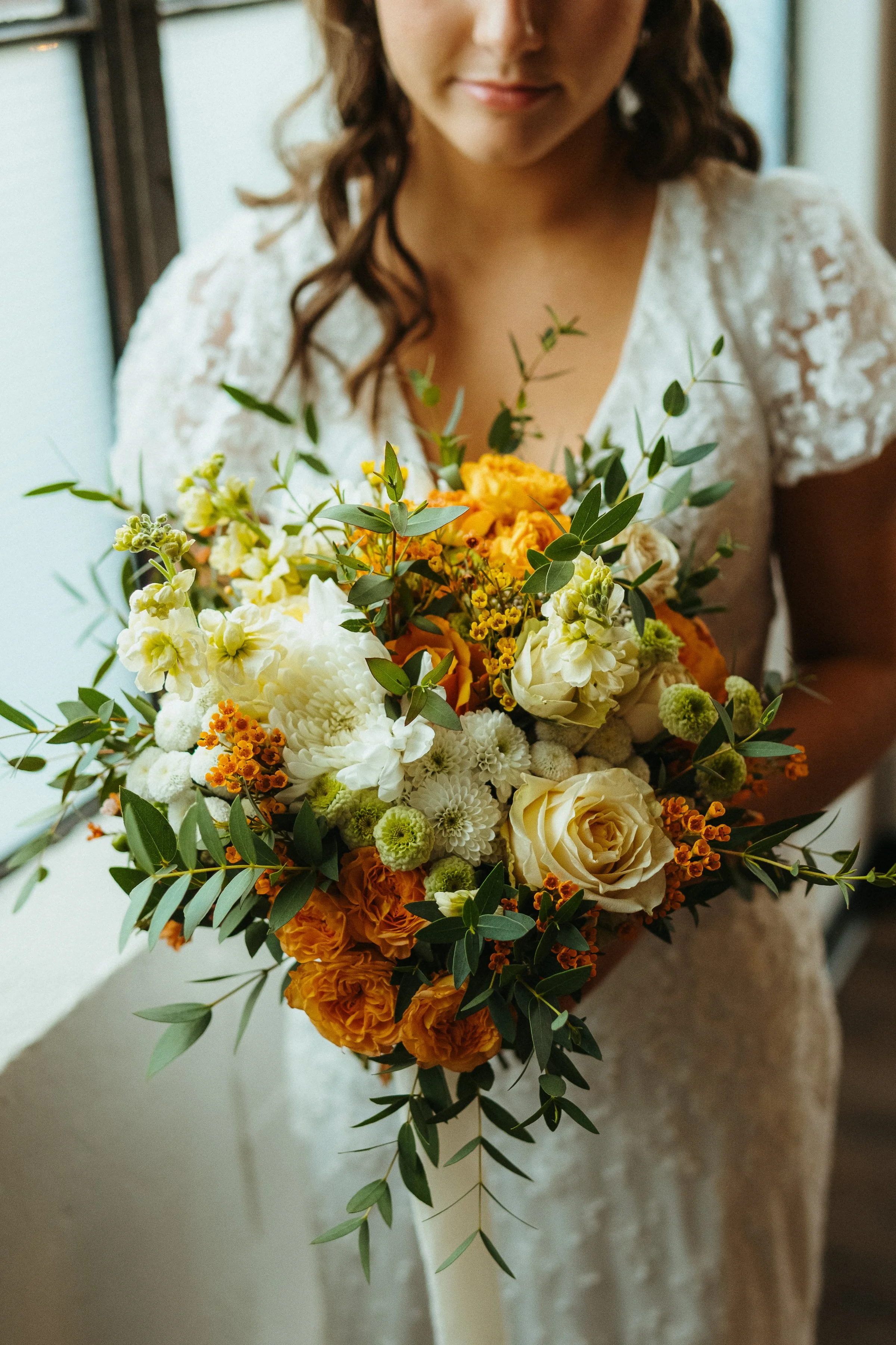 A woman in a lace dress holding a bouquet of white, orange, and yellow flowers with green foliage.