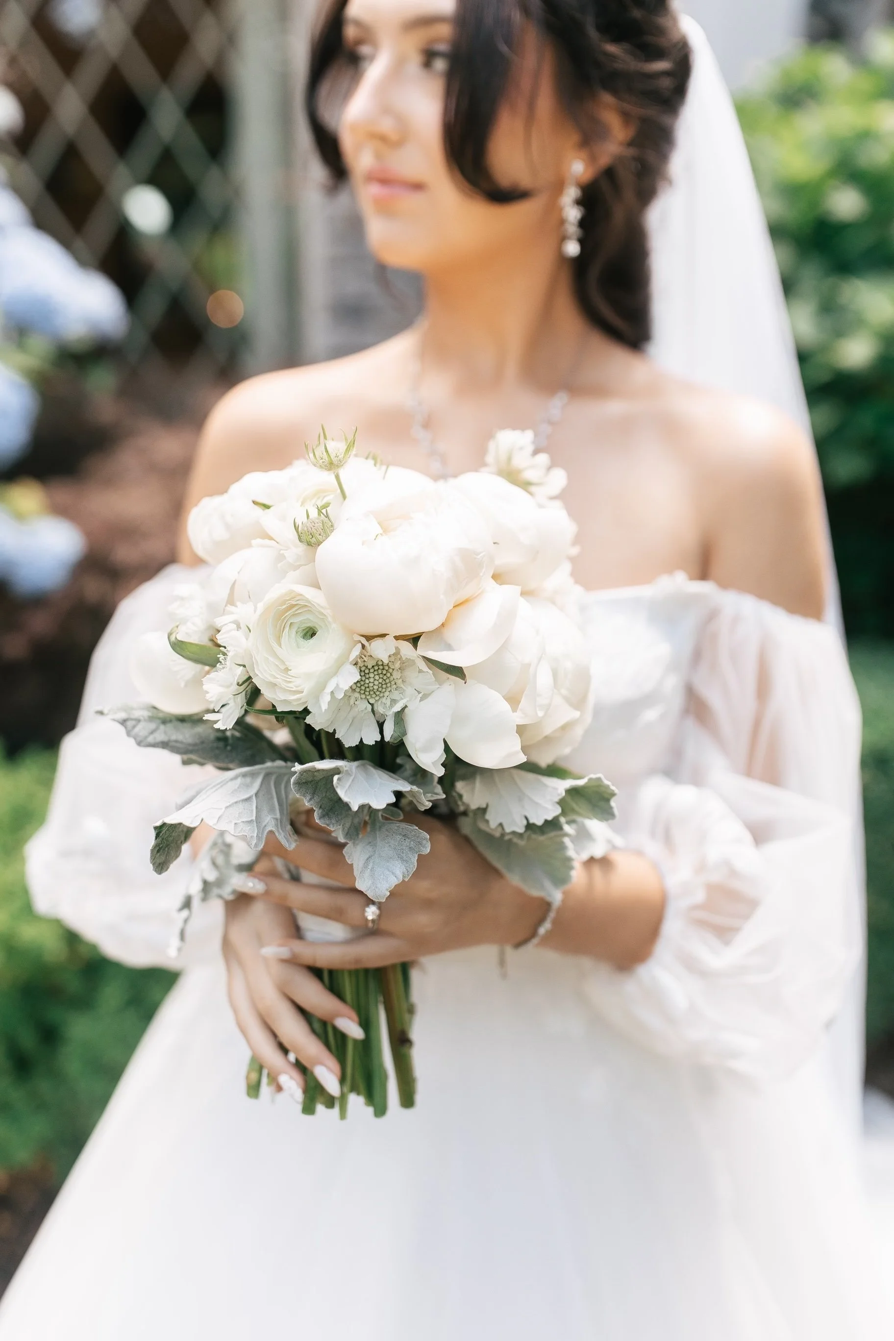 Bride holding a white floral bouquet outdoors, wearing an off-shoulder wedding dress with a veil and jewelry.