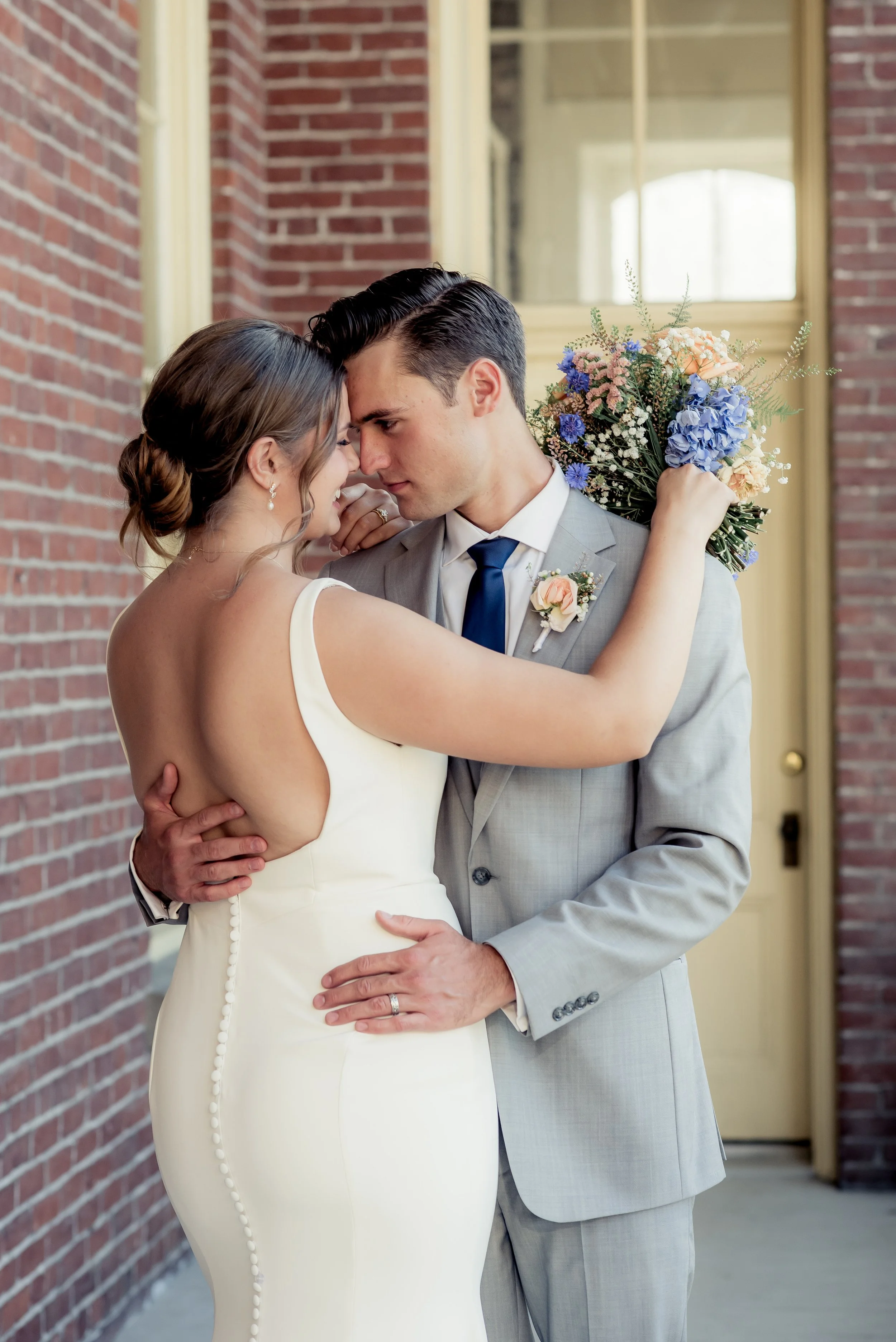 A bride and groom embrace closely, touching foreheads and gazing into each other's eyes outside a brick building, with the bride holding a bouquet of flowers.