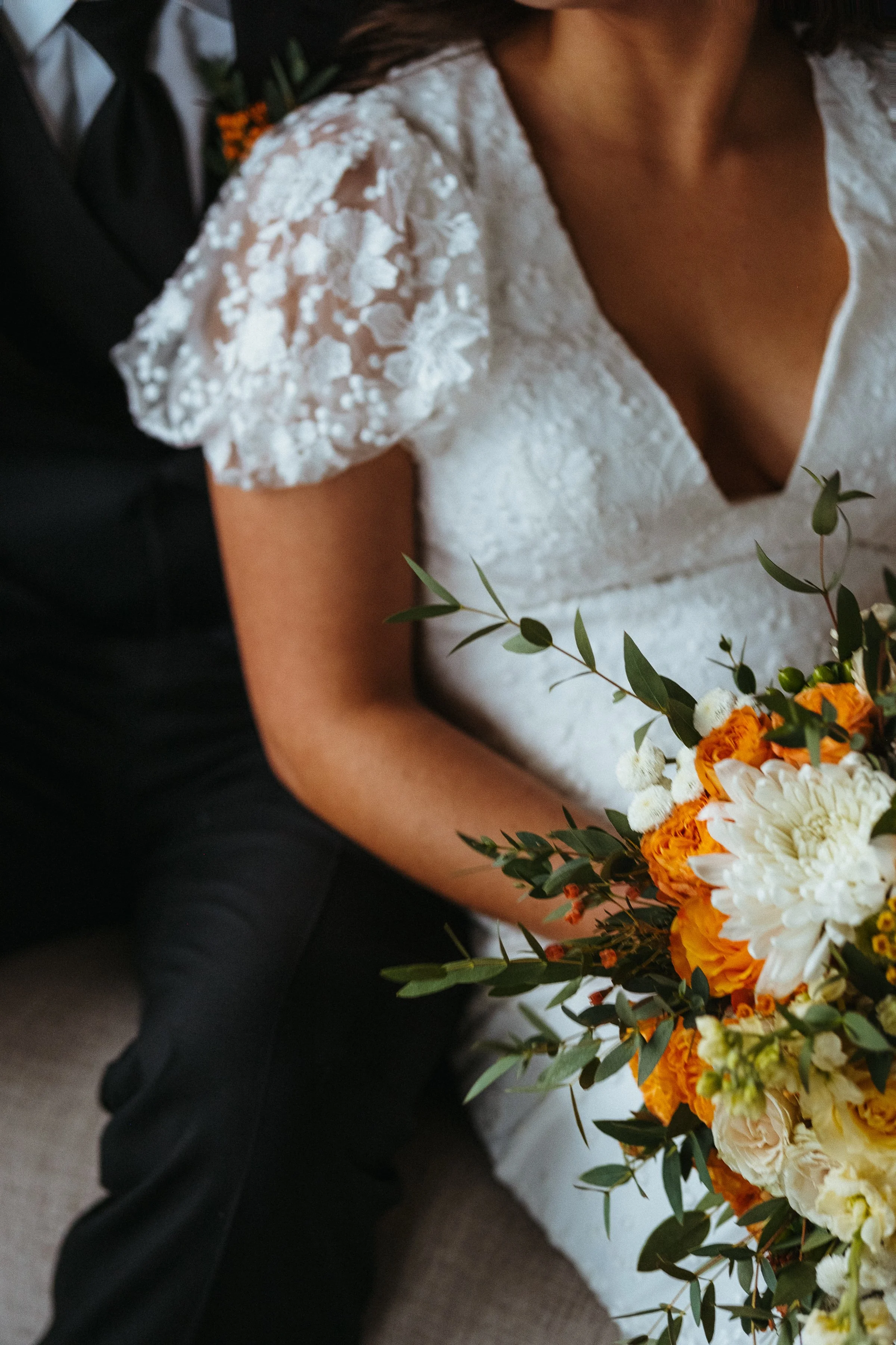 A woman in a white lace wedding dress holding a bouquet of orange, white, and yellow flowers with green foliage, sitting with a man in a black suit.