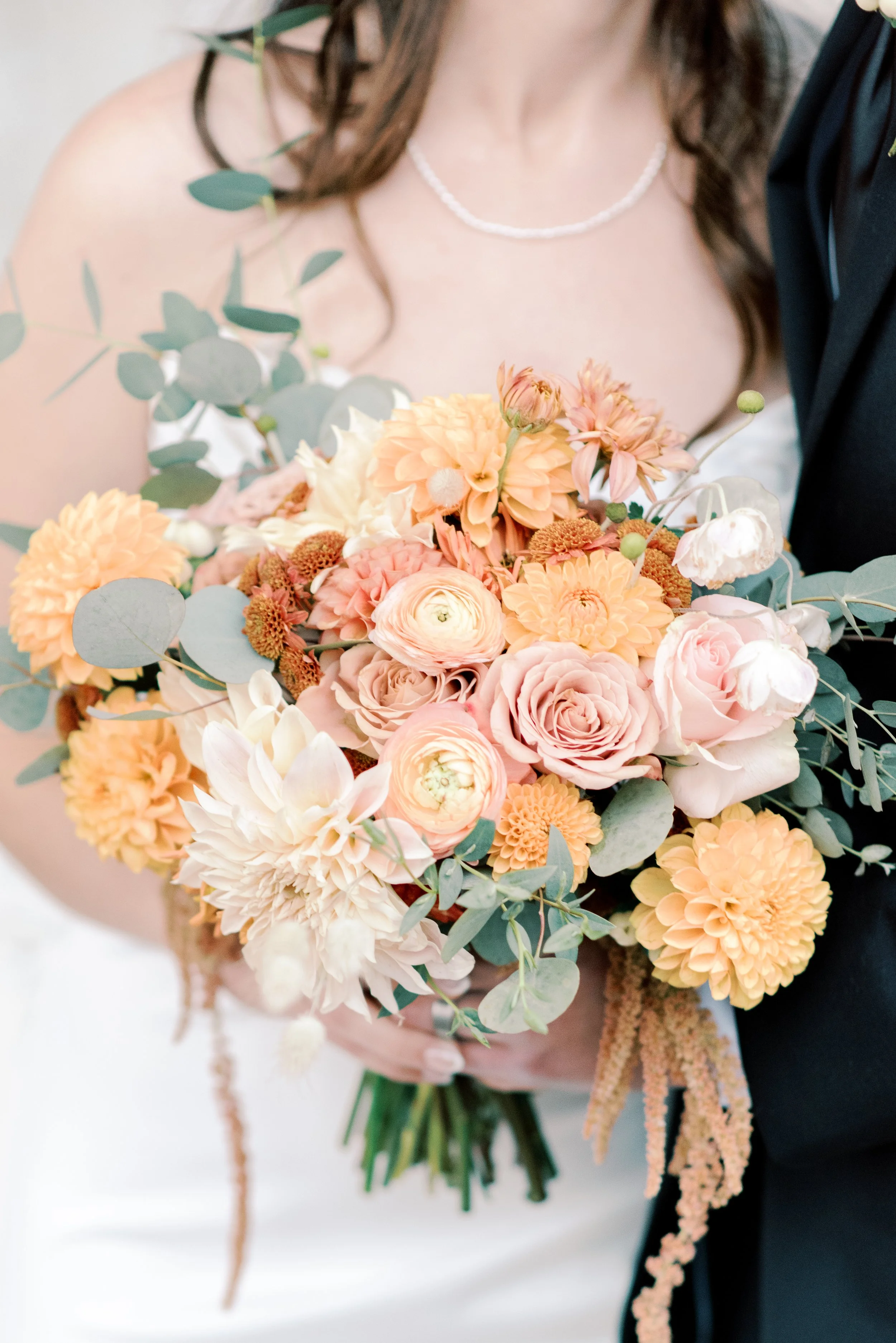 A bride holding a bouquet of peach, pink, and cream-colored flowers with greenery, near her wedding dress and a groom in a dark suit.