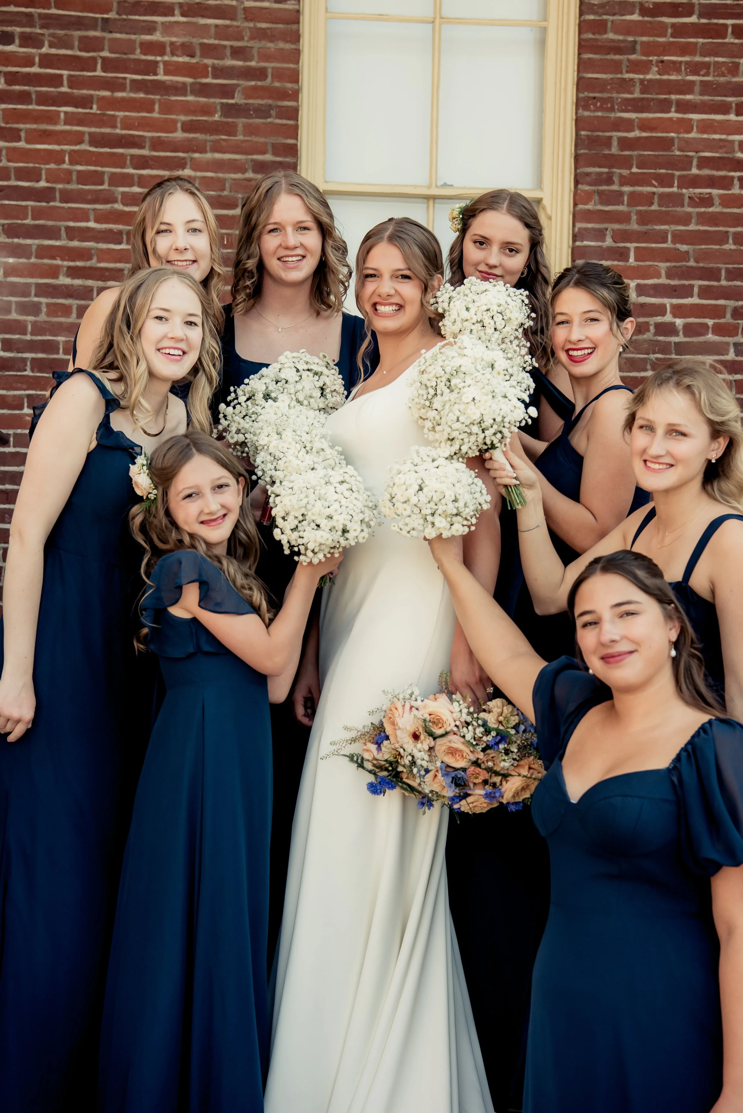 A bride in a white dress surrounded by women in navy blue dresses and a young girl in matching blue dress, holding bouquets of flowers, standing in front of a brick wall with a window.