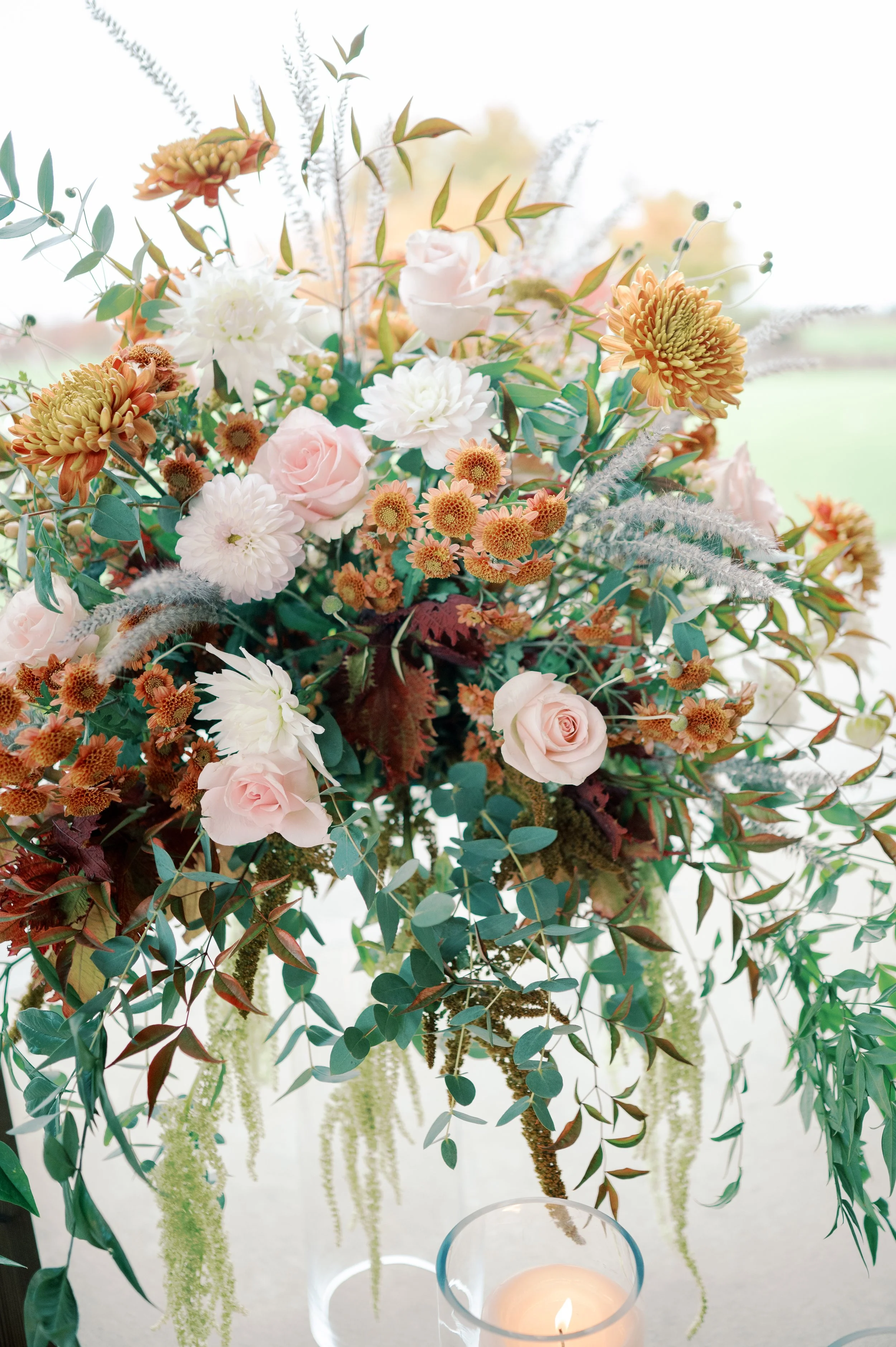 A large floral arrangement with pink roses, white dahlias, orange chrysanthemums, and various greenery and foliage, including eucalyptus leaves and trailing plants, displayed on a table with a lit candle in the foreground.