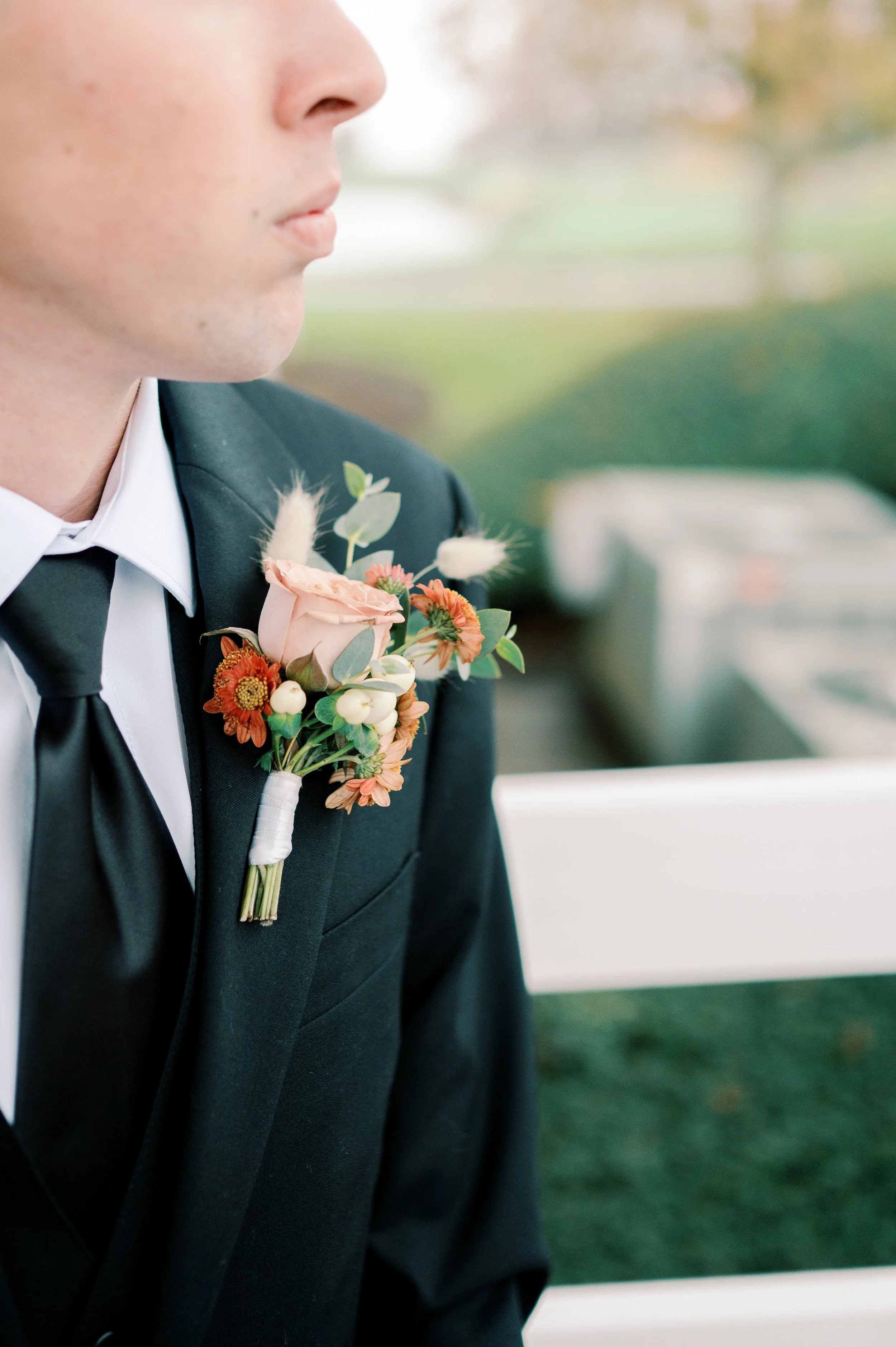 Close-up of a man in a black suit with a tie and a pink boutonniere on his lapel, showing part of his face and shoulder, outdoors with blurred green trees in the background.