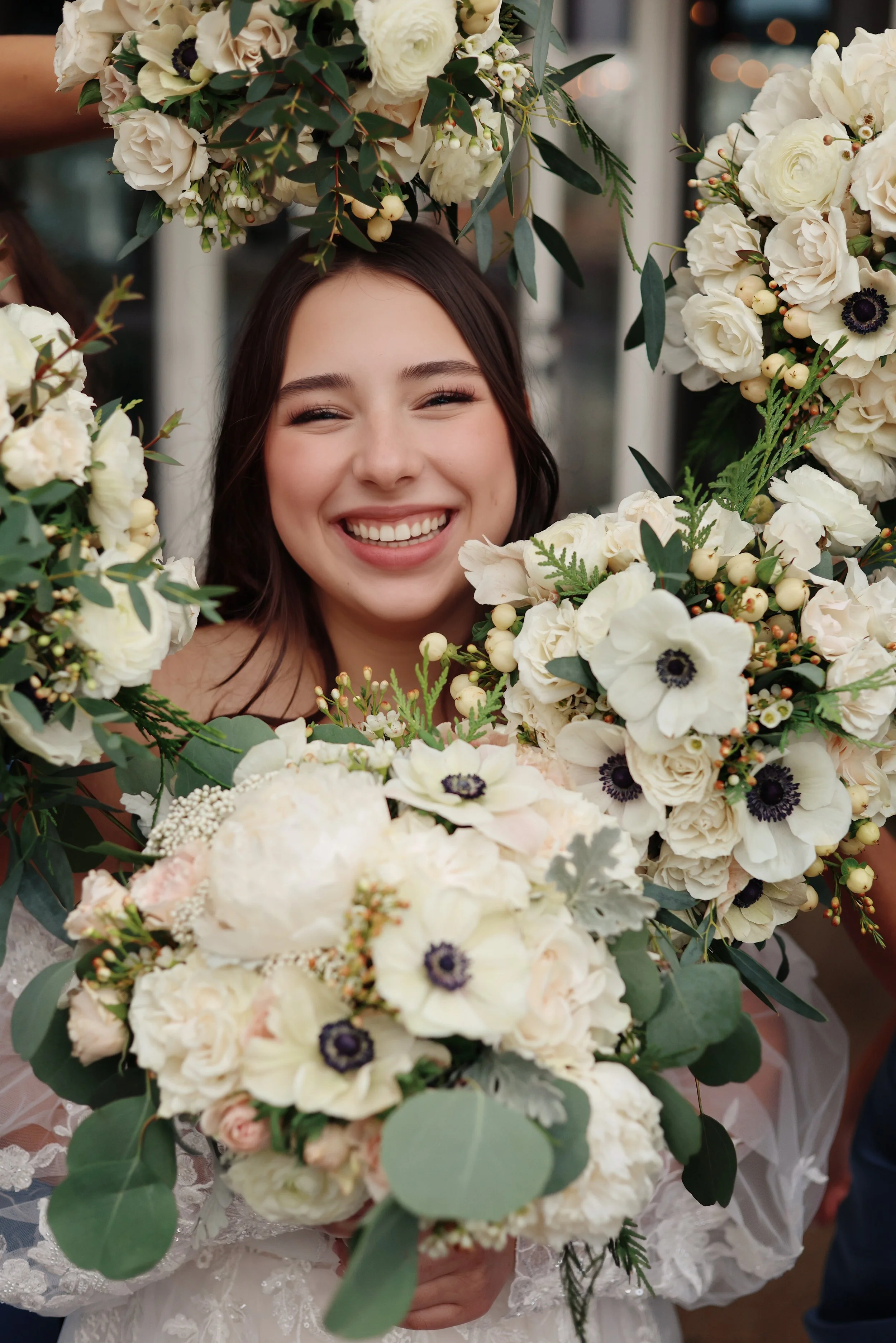 Smiling woman in wedding dress holding large bouquet of white flowers surrounded by additional white floral arrangements.