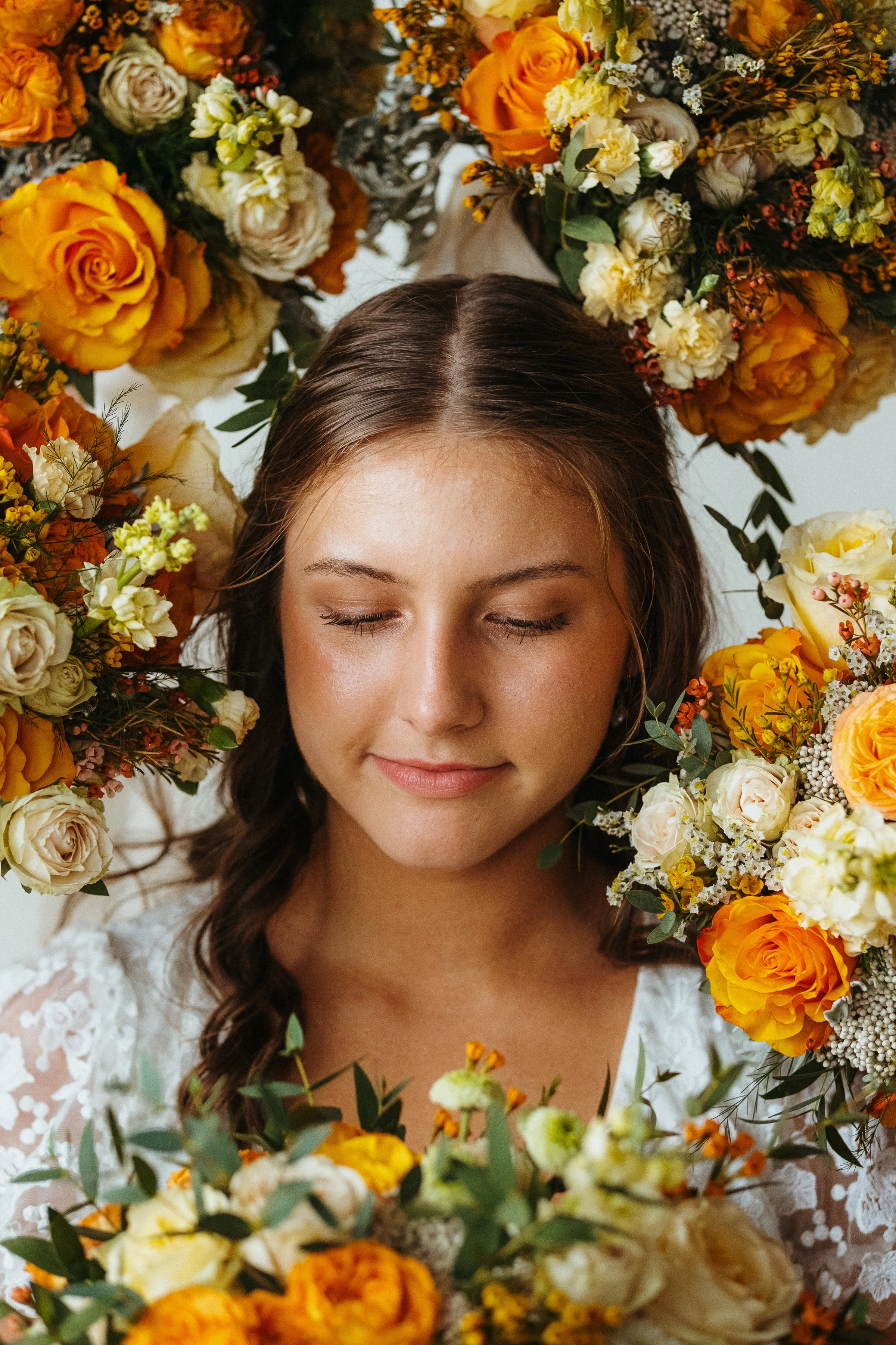 A young woman with wavy brown hair and closed eyes, surrounded by a large wreath of orange, yellow, and white flowers, wearing a white lace top.