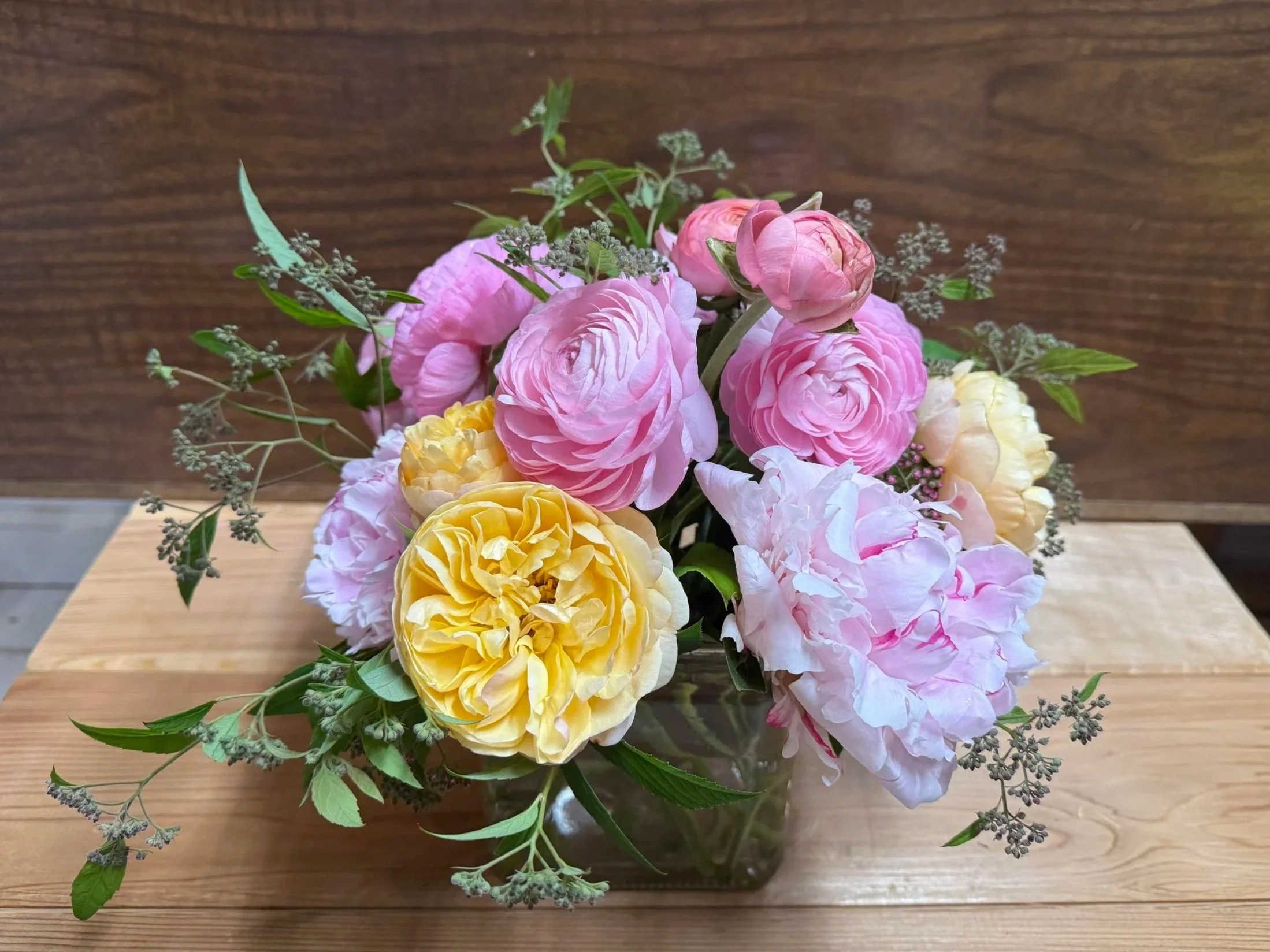 A glass vase with pink, yellow, and white peony flowers and green foliage on a wooden surface.