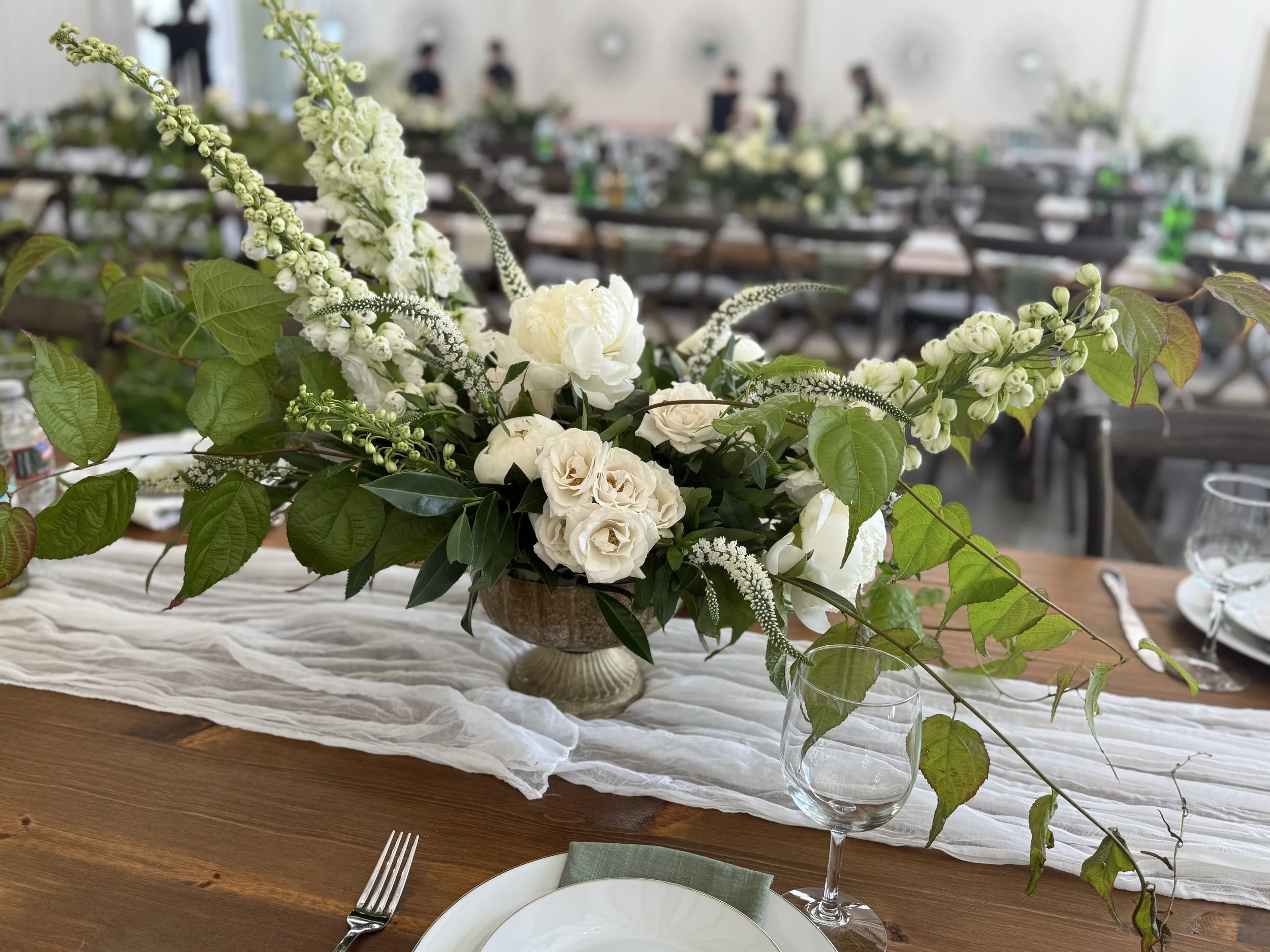 White floral centerpiece on a dining table at a social event, with tables, chairs, and people in the background.