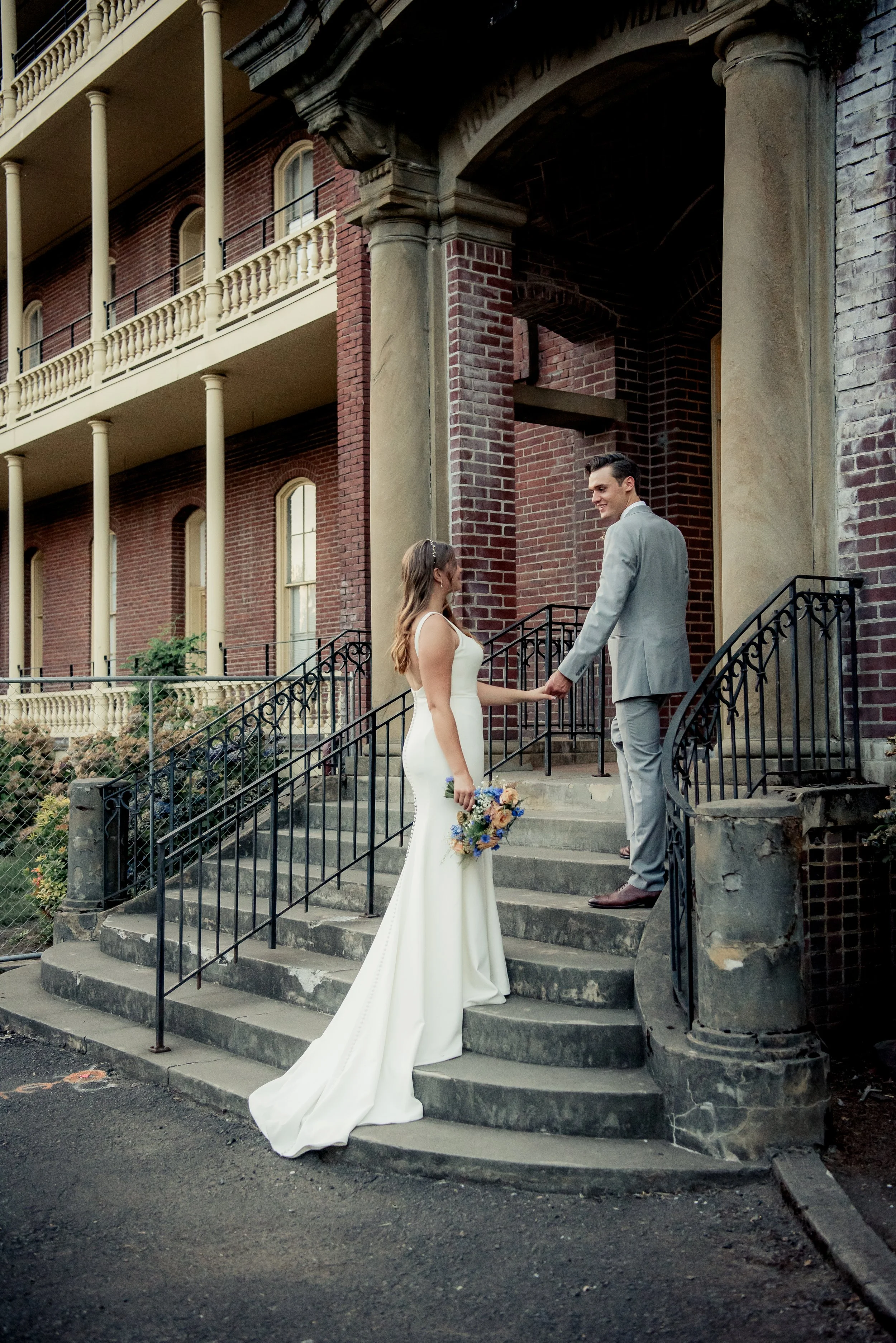 A bride and groom standing on the steps outside a historic brick building, holding hands and smiling at each other during their wedding.