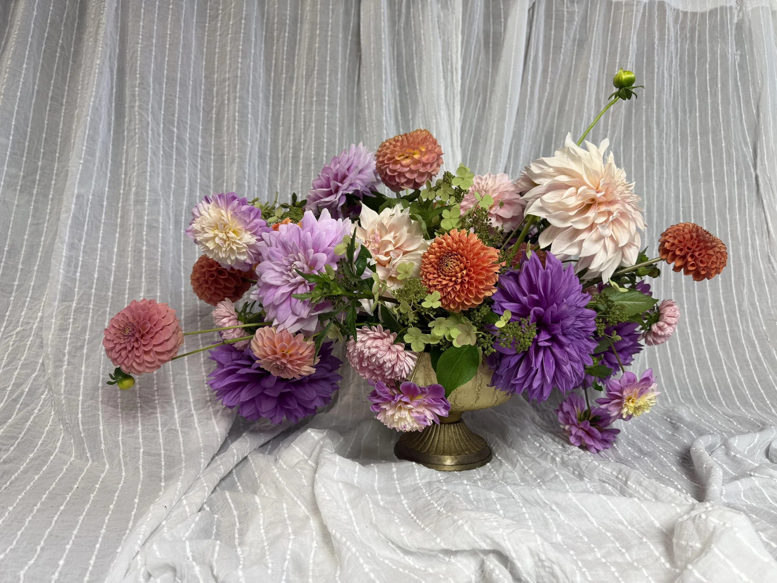 Colorful flower arrangement with pink, purple, orange, and white blooms in a gold vase against a light-colored fabric backdrop.