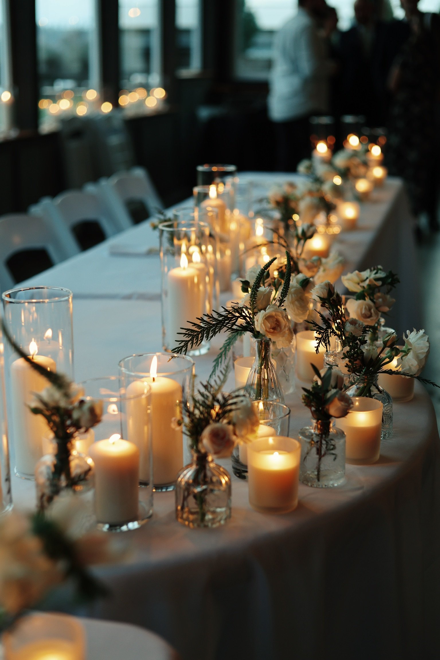 A long dining table decorated with lit candles in glass holders and small floral arrangements, set for a romantic evening or special event.