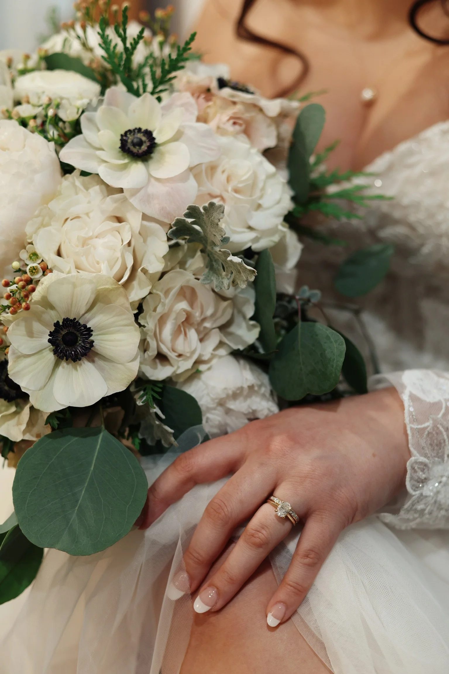 Close-up of a bride's hand with a wedding ring, resting on her lap with a wedding dress and lace sleeve visible, holding a bouquet of white roses, anemones, and greenery.