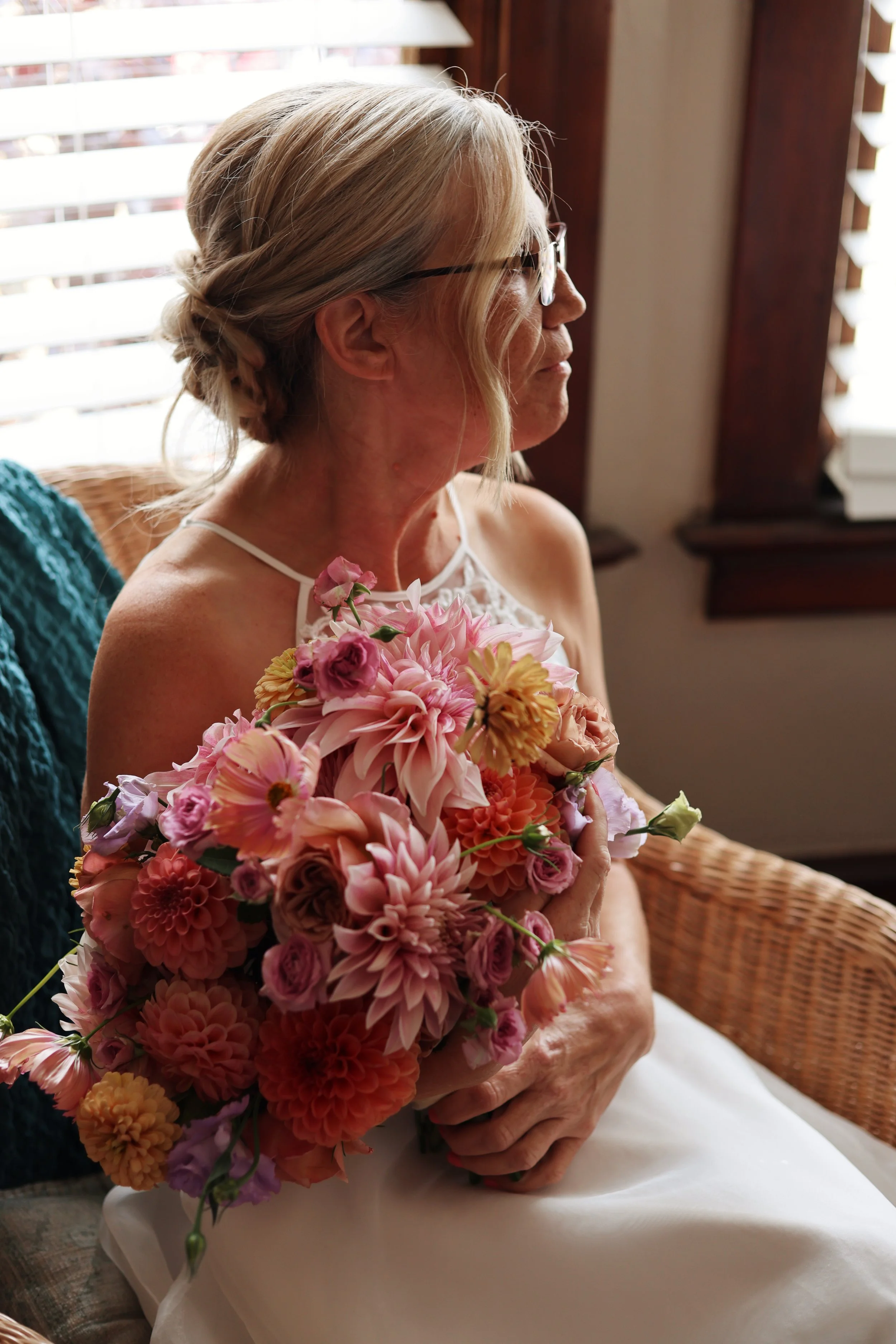 An elderly woman with glasses and gray hair styled in a bun, sitting indoors near a window with wooden blinds, holding a large bouquet of pink, peach, and purple flowers, wearing a white sleeveless dress.