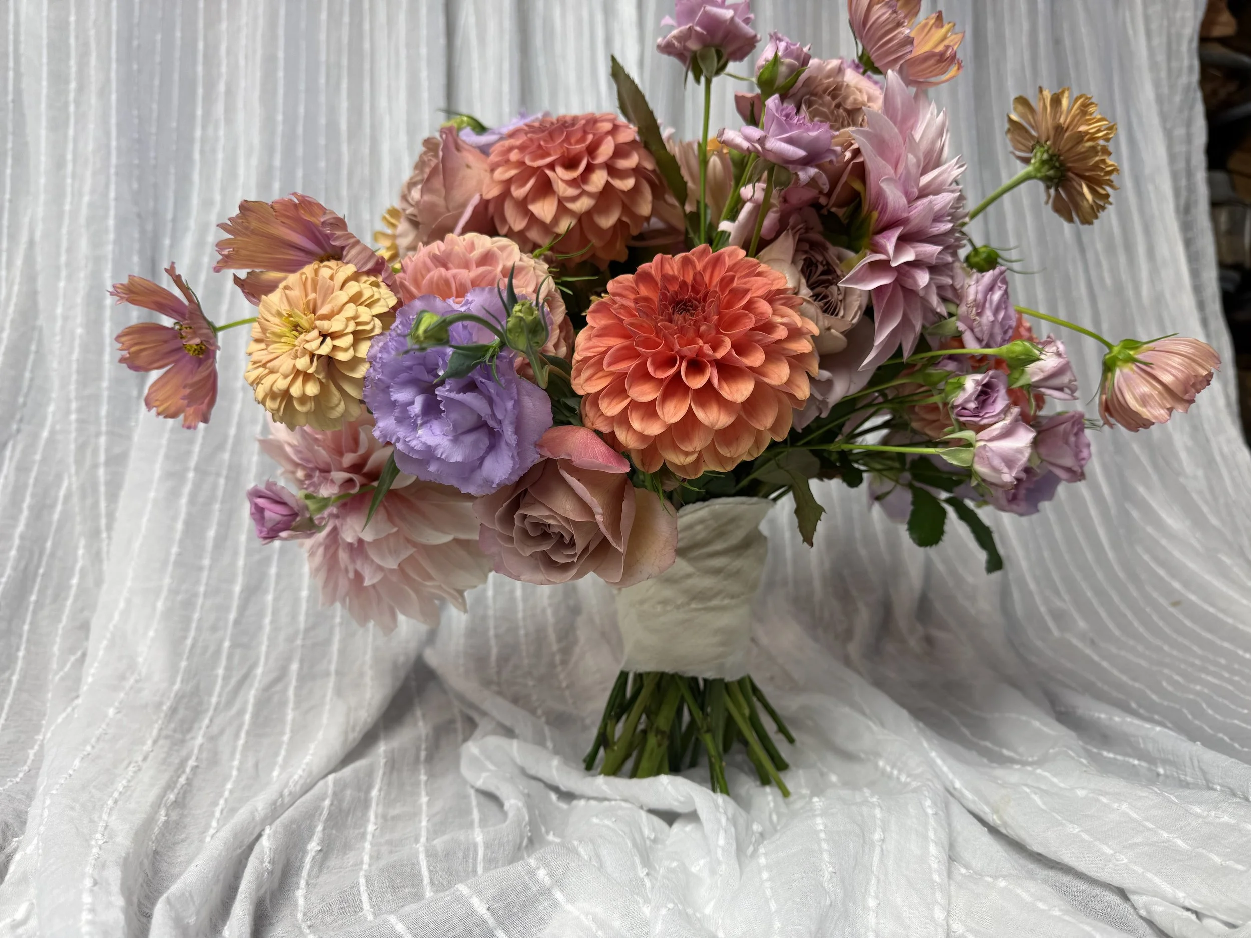 A bouquet of colorful flowers, including dahlias, roses, and lisianthus, arranged in a cream-colored vase, set against a white textured fabric background.
