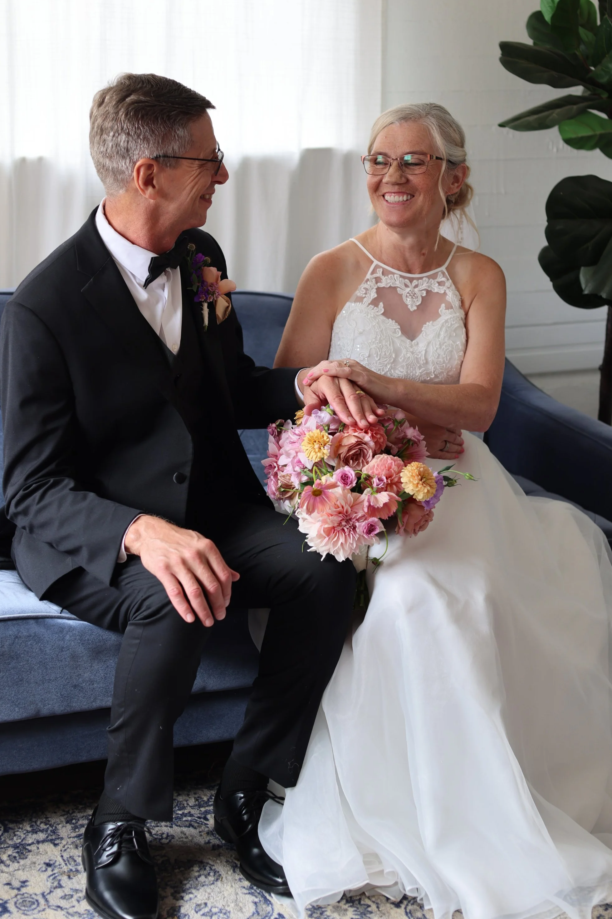 A bride and groom, both smiling and holding hands, sitting on a blue couch indoors. The bride is wearing a white wedding dress and glasses, holding a bouquet of pink and yellow flowers. The groom is in a black suit with a bow tie, glasses, and a small boutonniere.