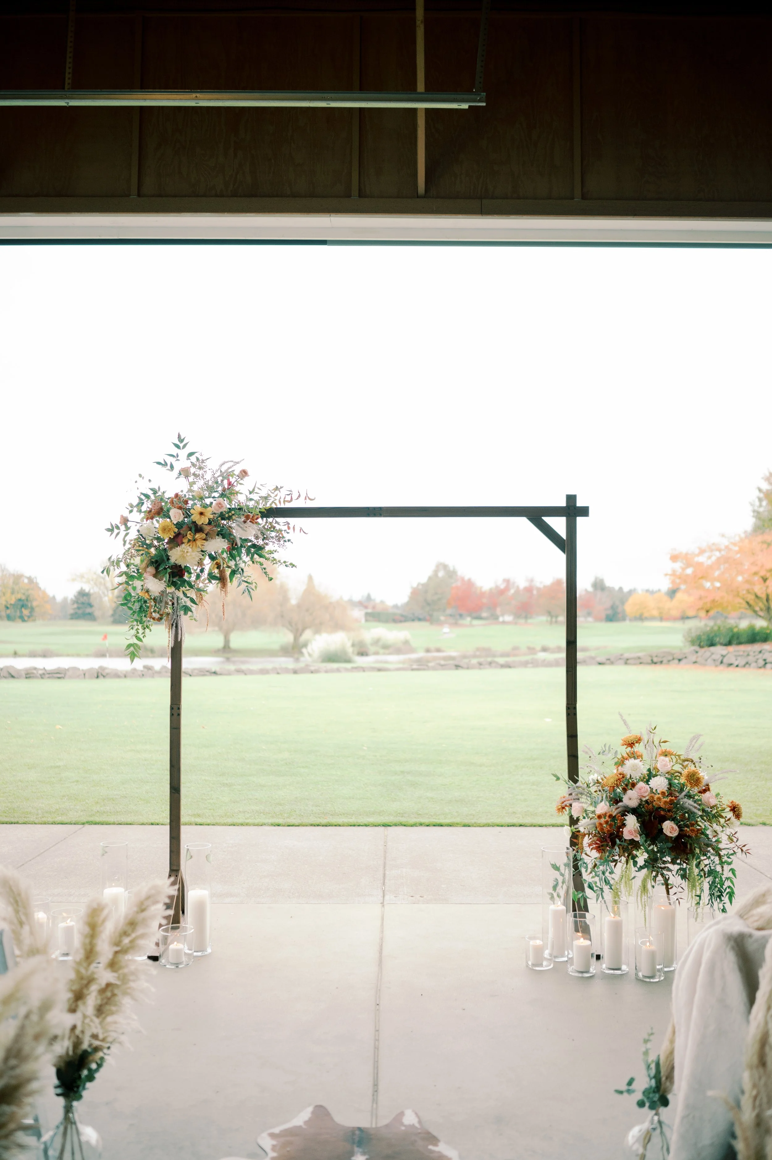 Wedding ceremony setup with a metal arch decorated with flowers and surrounded by candles outside near a golf course with trees in autumn.