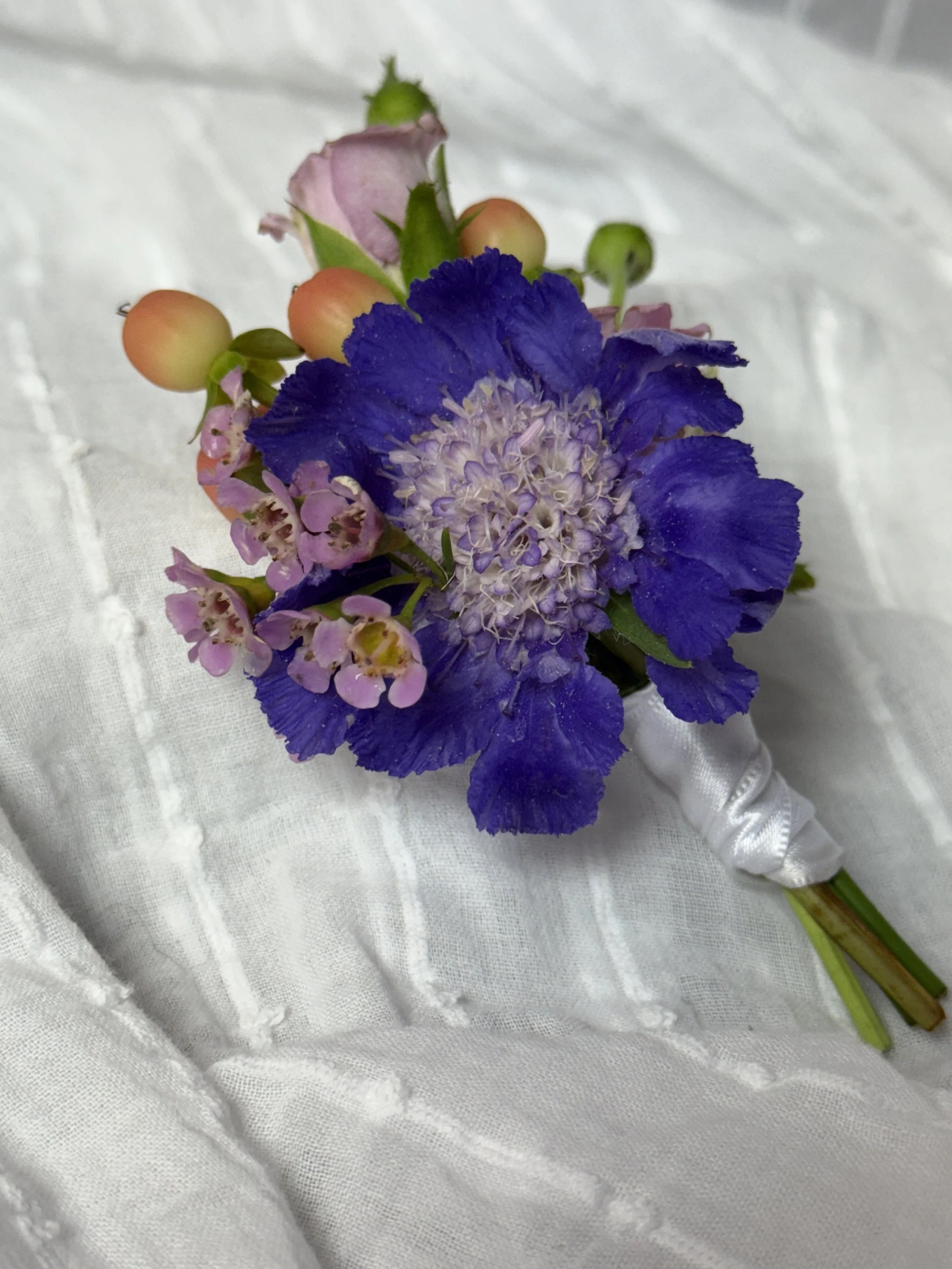 A close-up of a small bouquet of flowers with purple, pink, and cream-colored blooms, wrapped with white satin ribbon, resting on a white textured fabric background.