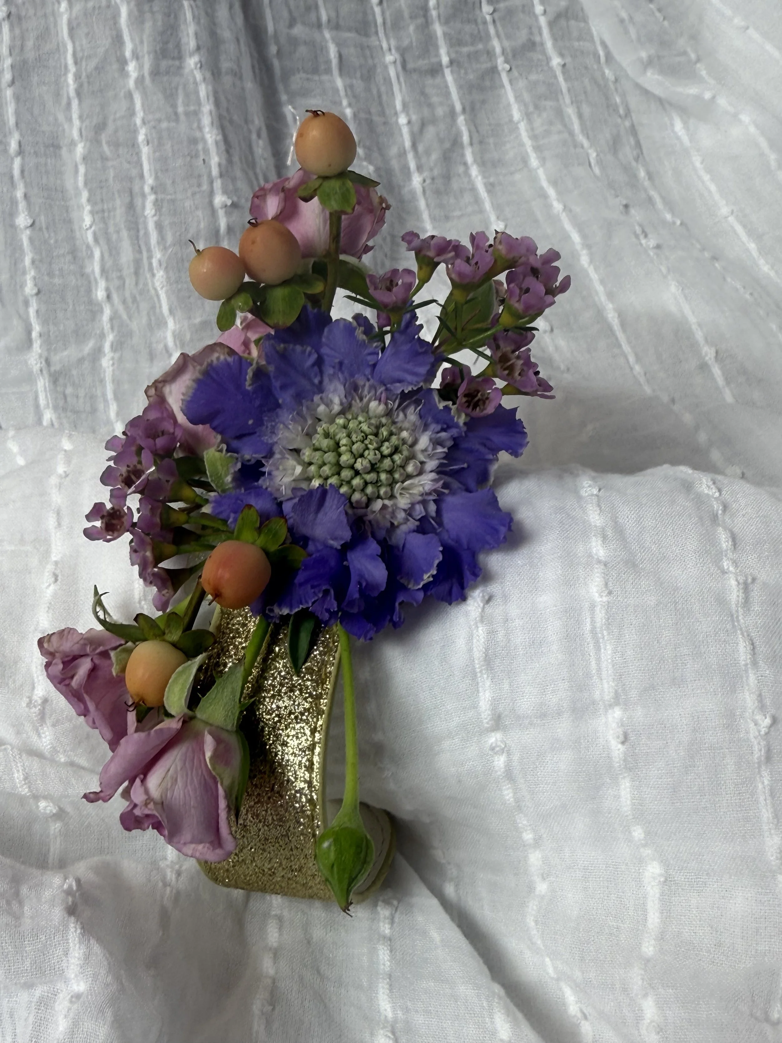 A small floral arrangement in a gold-colored vase featuring purple, pink, and green flowers with pale berries, set on white fabric with embroidered details.