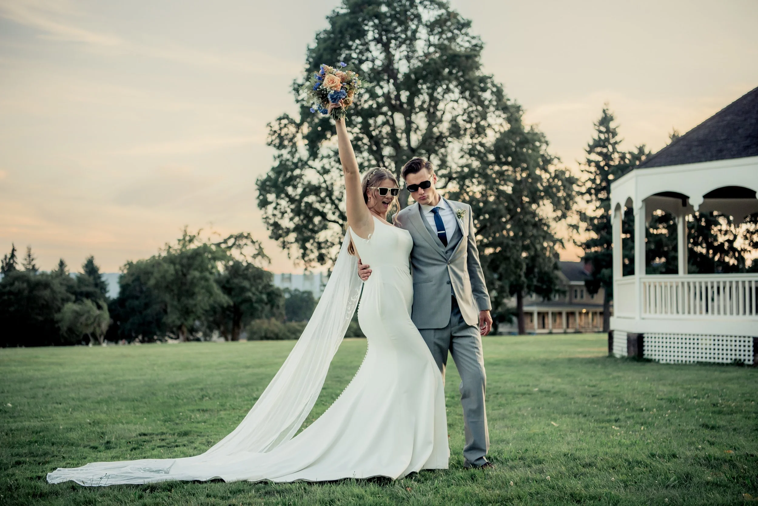 Bride and groom celebrating outdoors at sunset, with the bride holding a bouquet and wearing sunglasses, in a grassy park.