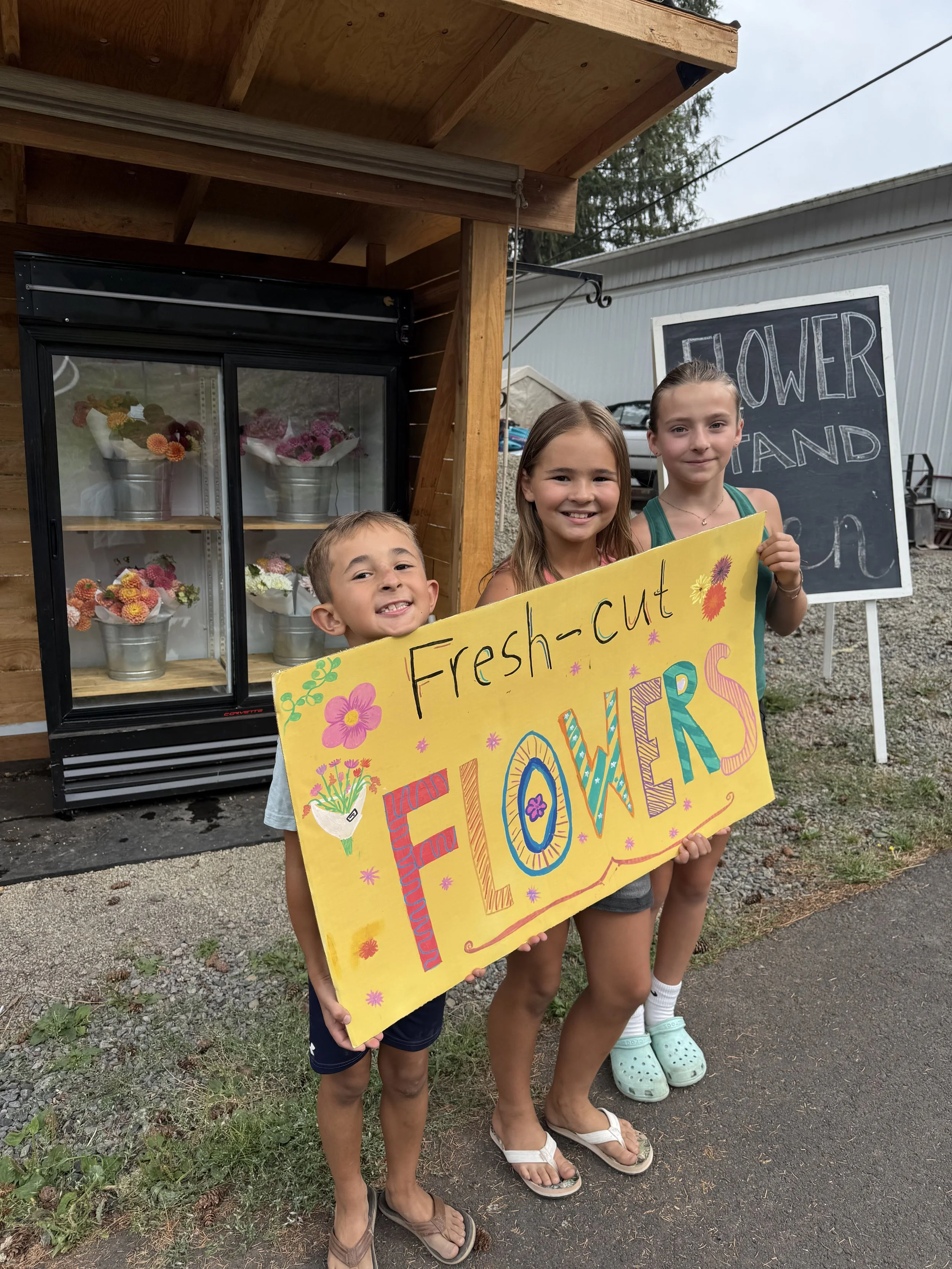 Three children holding a colorful sign that reads "Fresh-cut FLOWERS" outside a flower stand with a refrigerator displaying flowers and a chalkboard sign in the background.