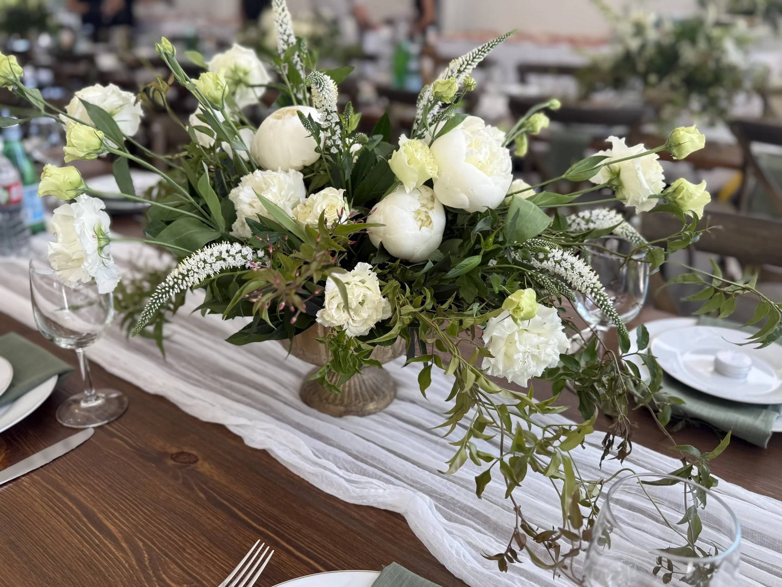 Elegant floral centerpiece with white peonies, roses, and greenery on a wooden table with a white table runner for a formal event.