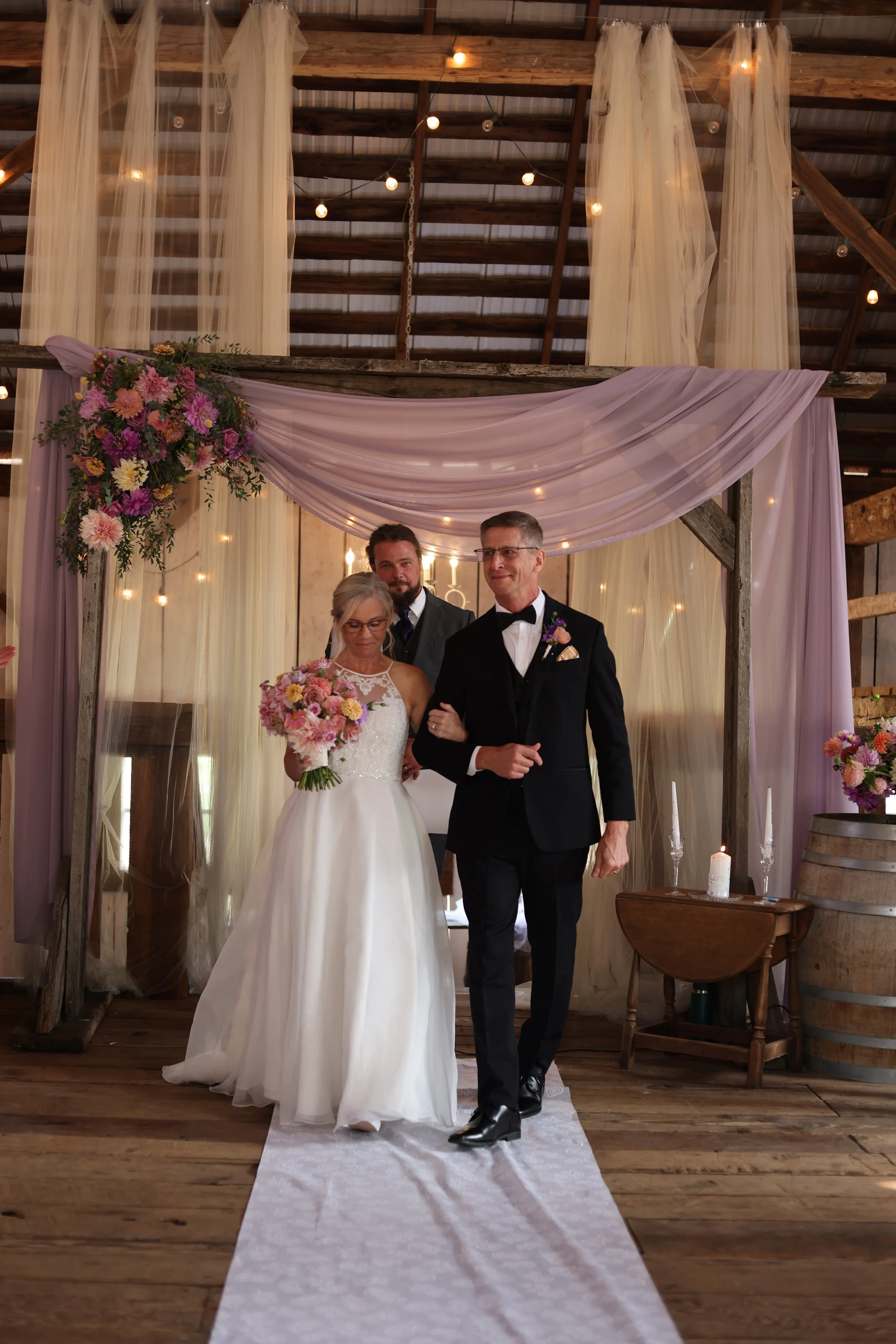 A wedding ceremony with a bride in a white wedding dress holding a bouquet, a groom in a black tuxedo, and a officiant standing behind them in a rustic indoor setting decorated with purple and pink flowers, draped fabric, and warm string lights.