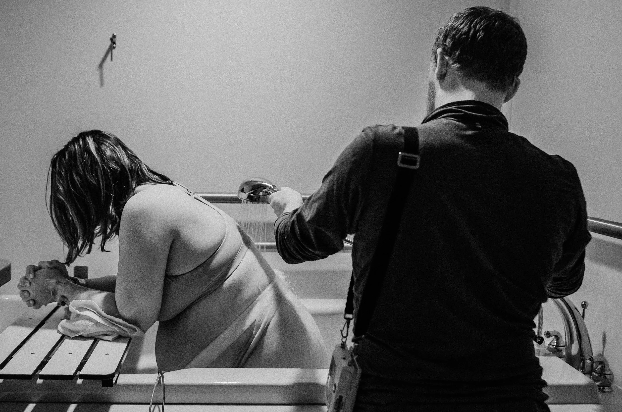 A woman leaning over a hospital tub, her family member or caregiver is assisting her, in a grayscale photo.