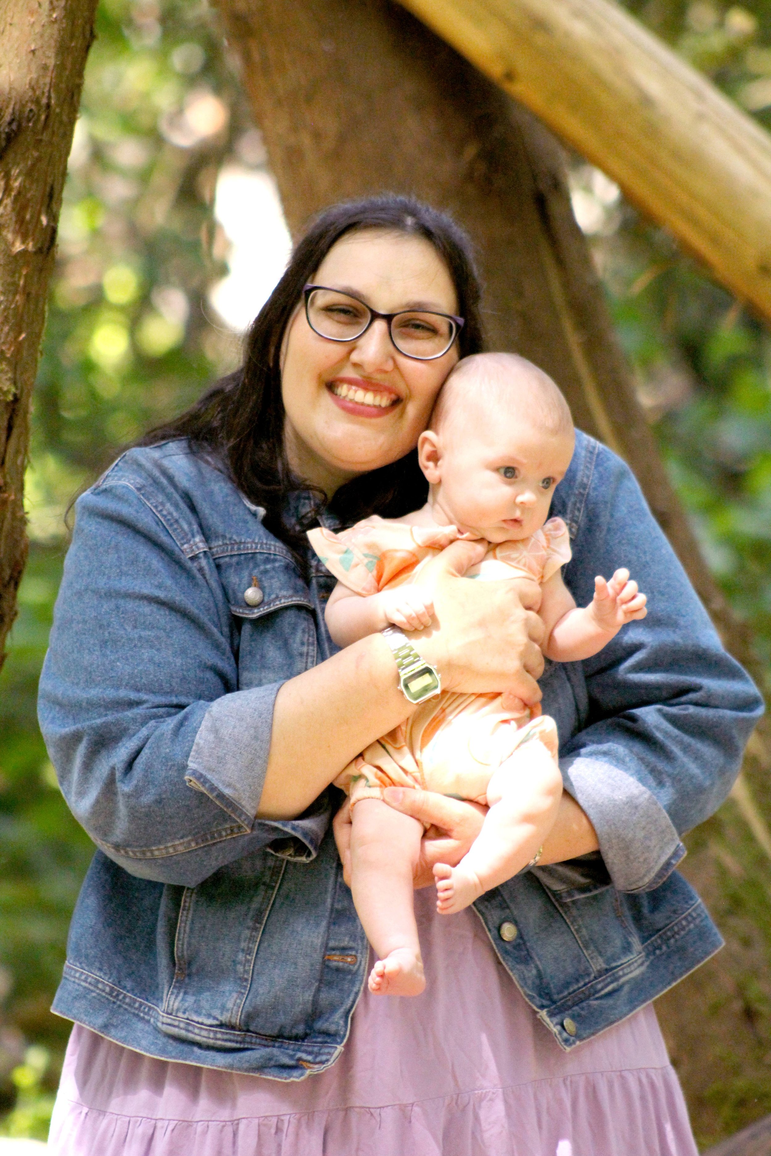 A woman with dark hair, glasses, and a denim jacket holding a baby girl outdoors in a wooded area.