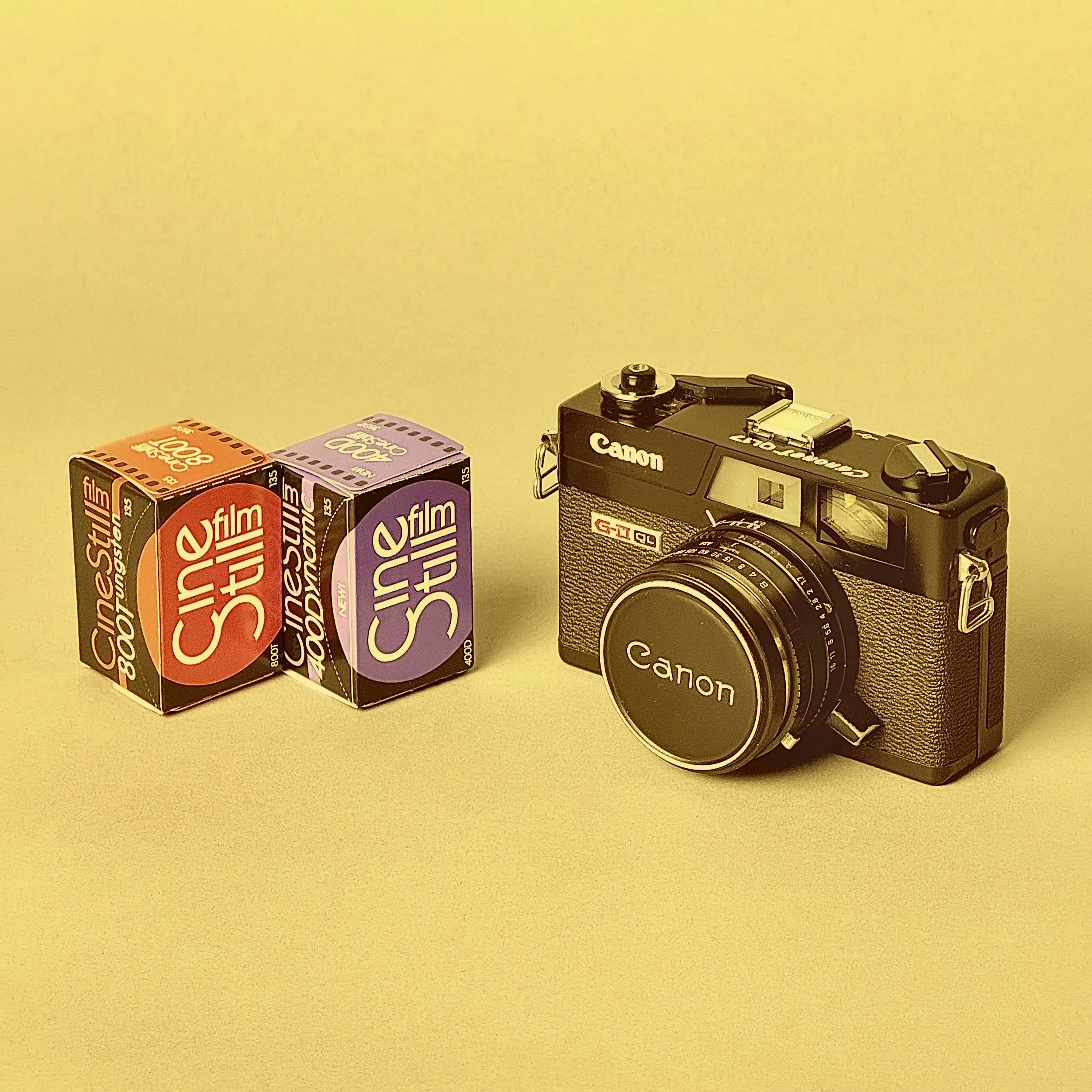A vintage Canon film camera with a lens cap, and two boxes of film, one labeled CineStill 800T and the other CineStill 50D, on a plain surface against a yellow background.