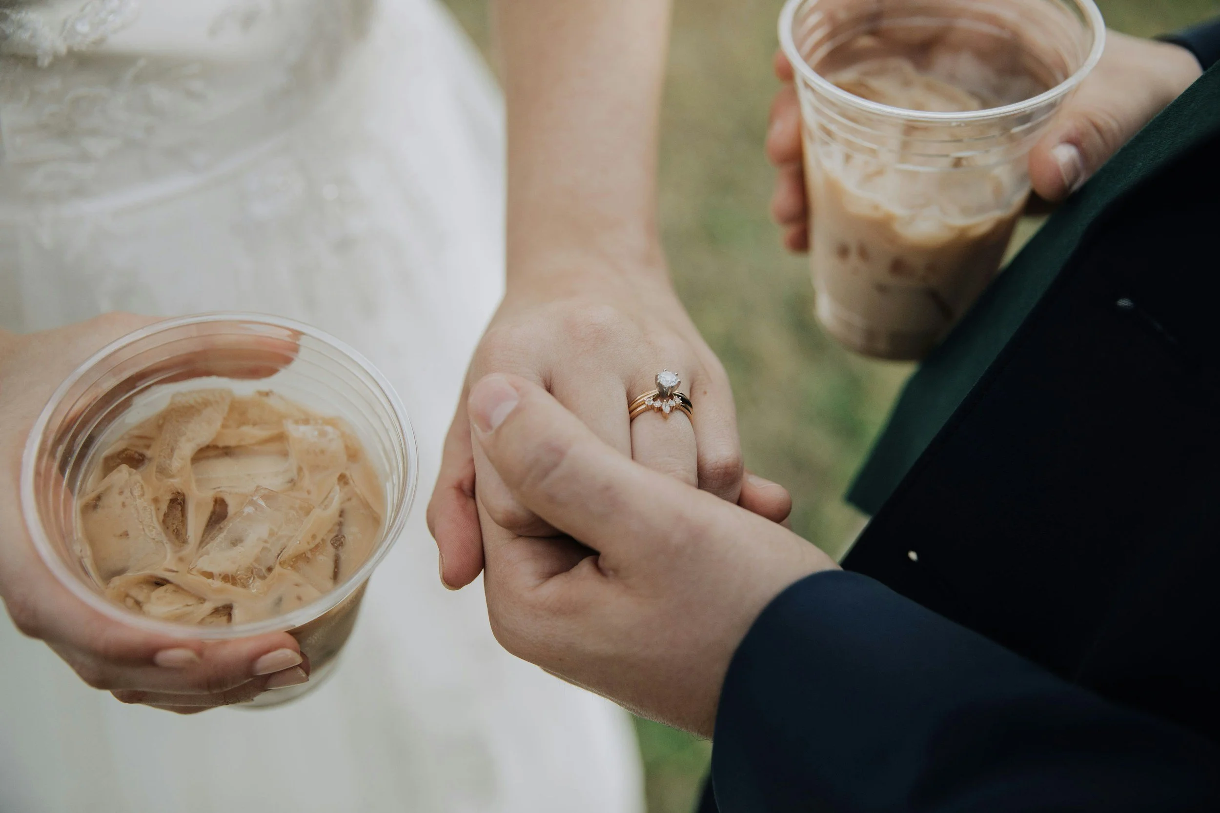 A bride and groom holding hands, with the bride wearing an engagement ring and the groom in a tuxedo, each holding a plastic cup of iced coffee.