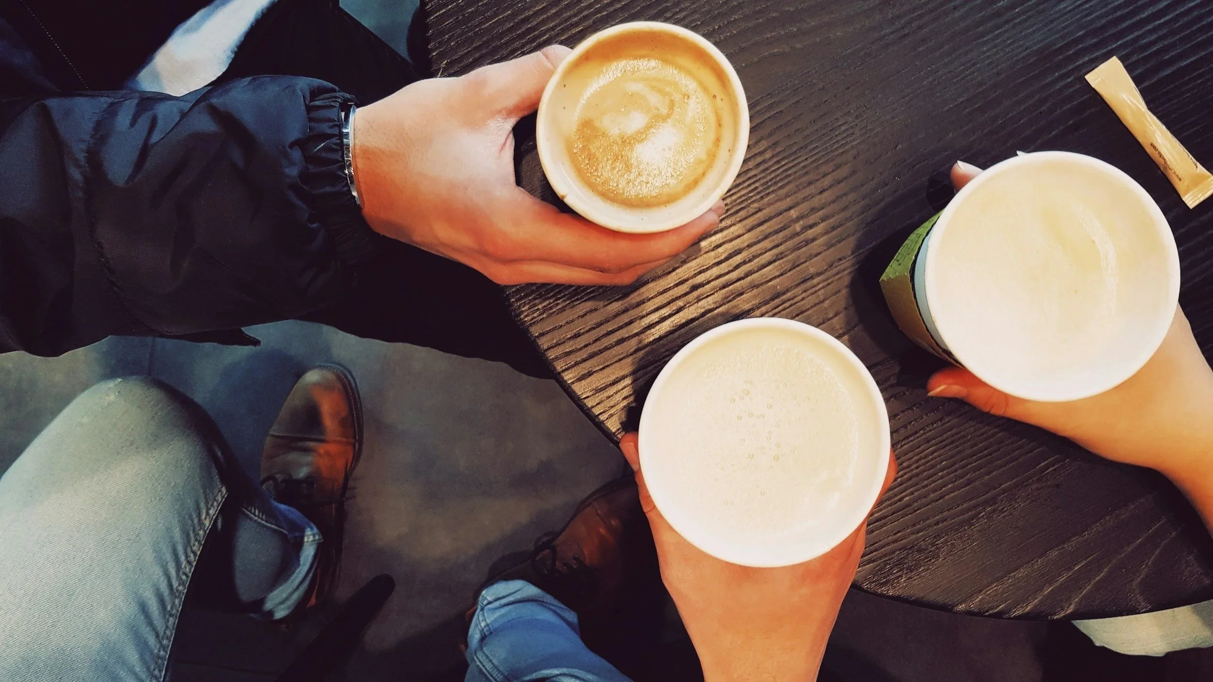 Three people holding cups of coffee or tea above a black wooden table, with snacks on the side.