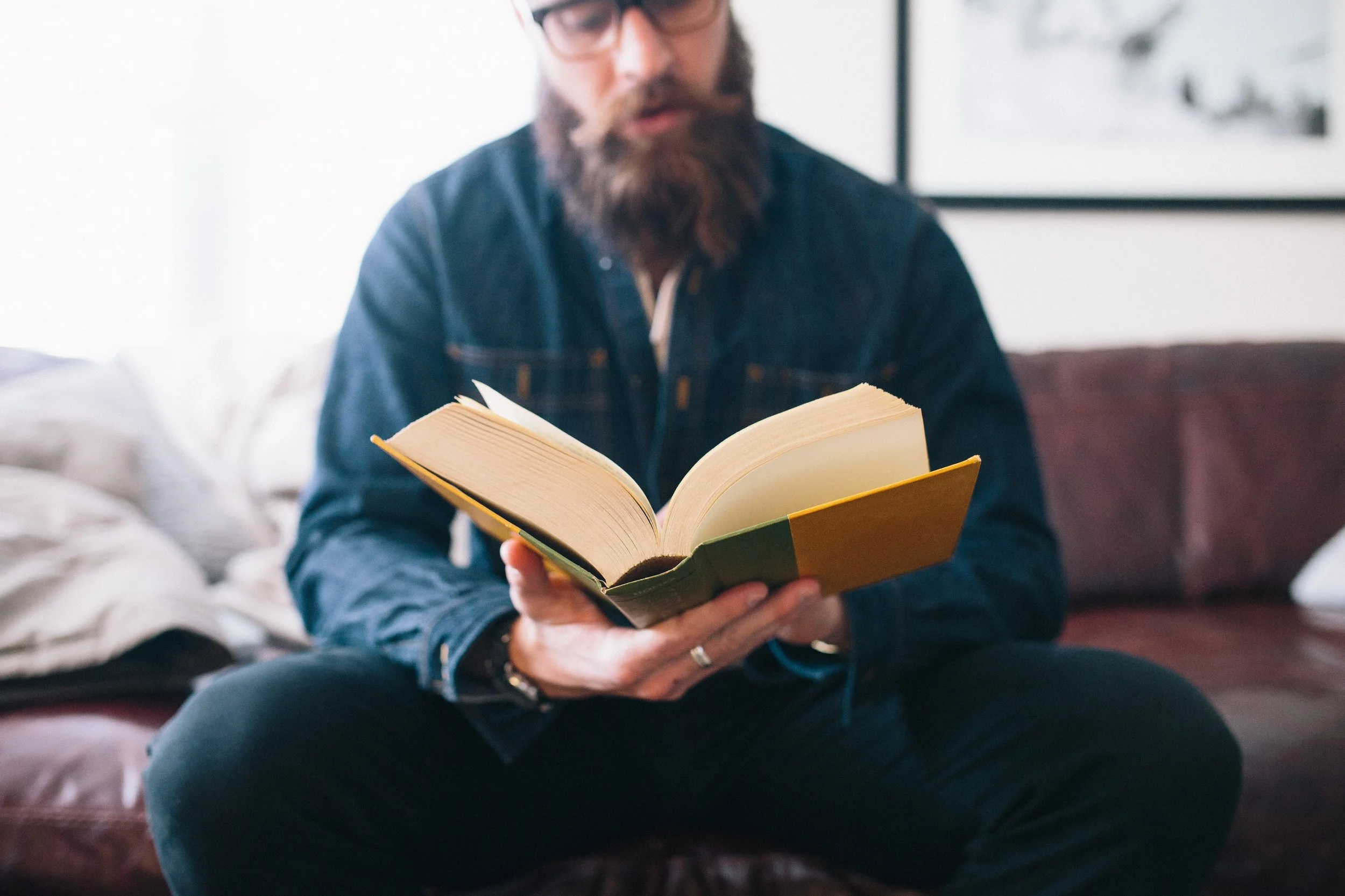 A man with glasses and a beard sitting on a brown leather sofa, reading a hardcover book.