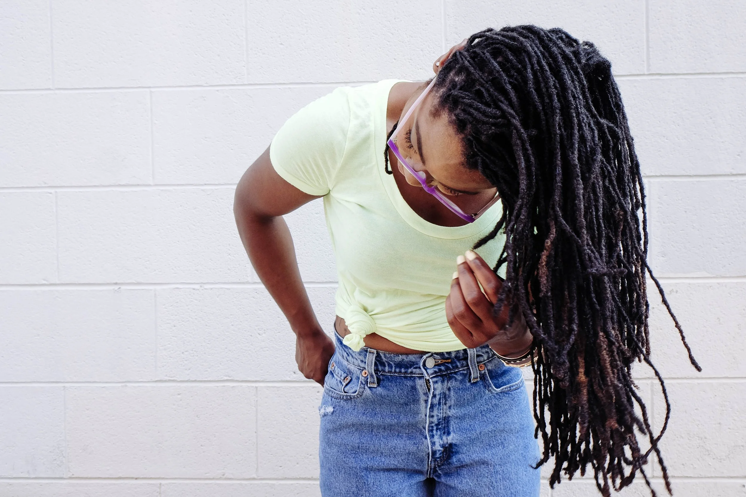 A woman with long dreadlocks, wearing a light yellow T-shirt and blue jeans, looking down and holding her stomach, standing against a white brick wall.