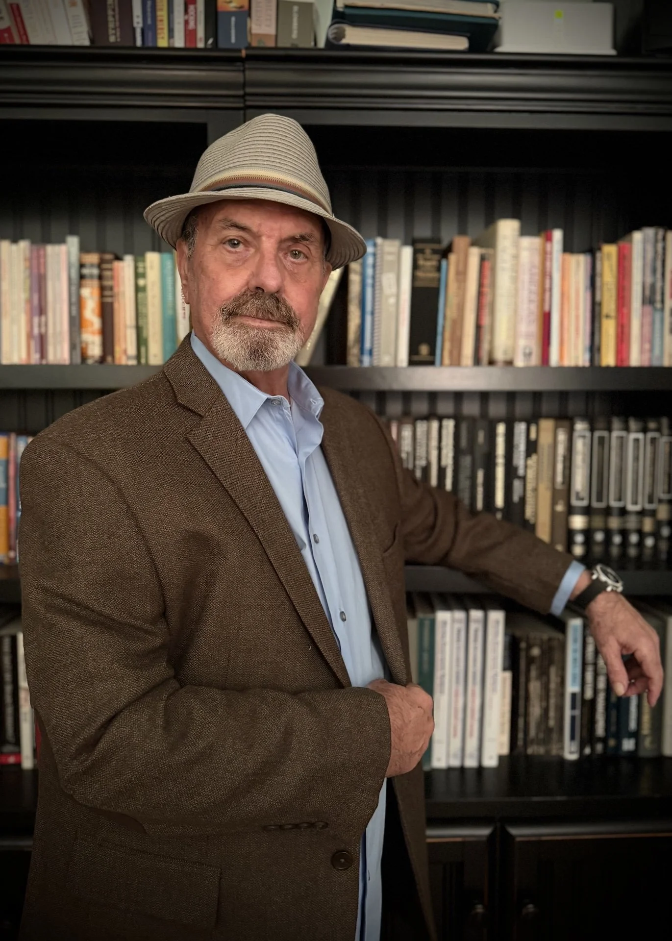 A man with a beard wearing a fedora hat, brown blazer, and light blue shirt standing in front of black bookshelves filled with books.