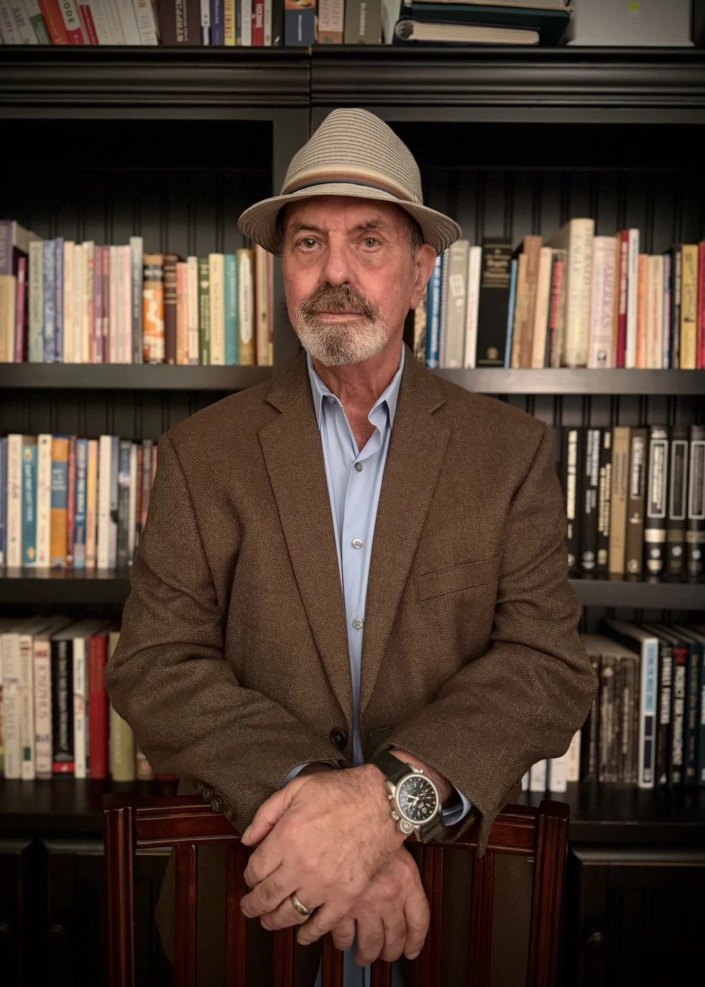 Steve Osborne, PhD, Beyond Chance with a beard and mustache wearing a beige fedora, a brown blazer, and a light blue shirt. He is standing in front of a bookshelf filled with books, leaning on a wooden chair with his hands clasped.