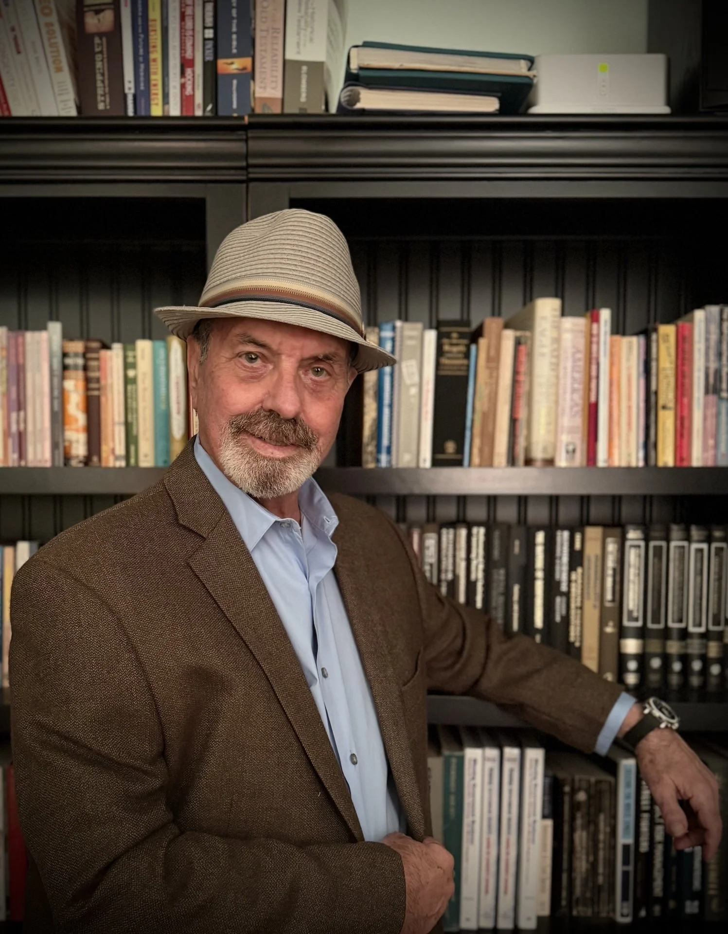 A man with a beard and mustache wearing a light-colored fedora, brown blazer, and light blue shirt, standing in front of a bookshelf filled with books. He is smiling and touching a book on the lower shelf with his right hand.