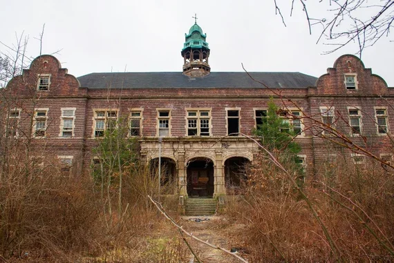 An old, abandoned brick building with a central facade, arched entrance, and a tower with a green dome on top, surrounded by overgrown bushes and trees.
