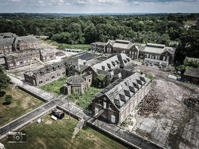 Aerial view of an abandoned, partially ruined industrial or institutional complex surrounded by trees and open fields.
