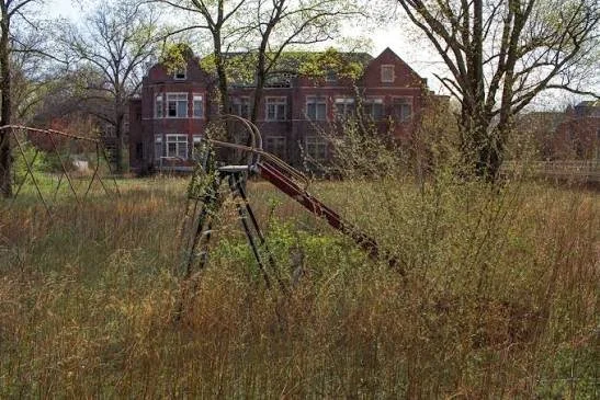Old, abandoned playground slide with overgrown grass and bushes in front of a large, historic brick building surrounded by trees.