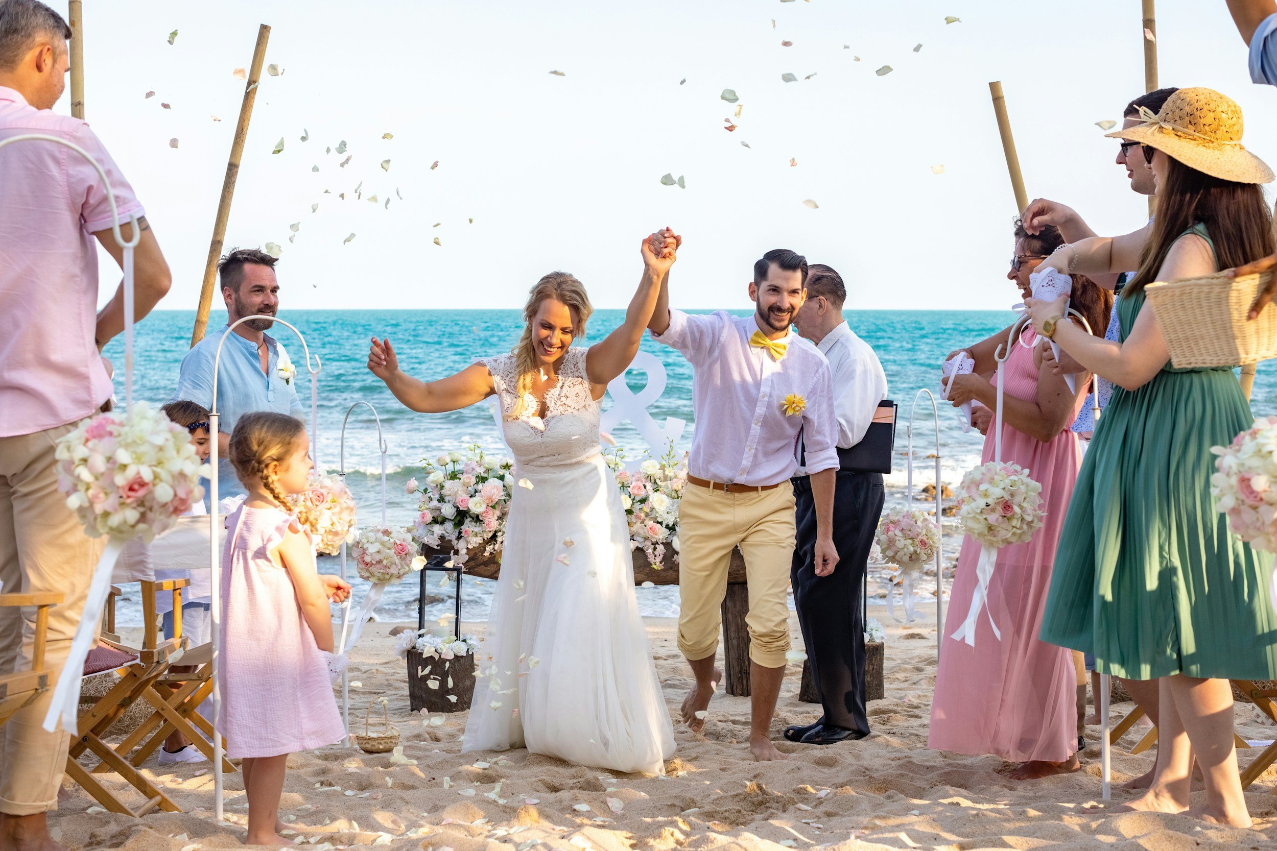 A beach wedding ceremony with a bride and groom holding hands, smiling, and walking barefoot on the sand. They are surrounded by friends and family, some of whom are wearing summer dresses and hats. The ocean is visible in the background with party confetti in the air. The scene is bright and cheerful.
