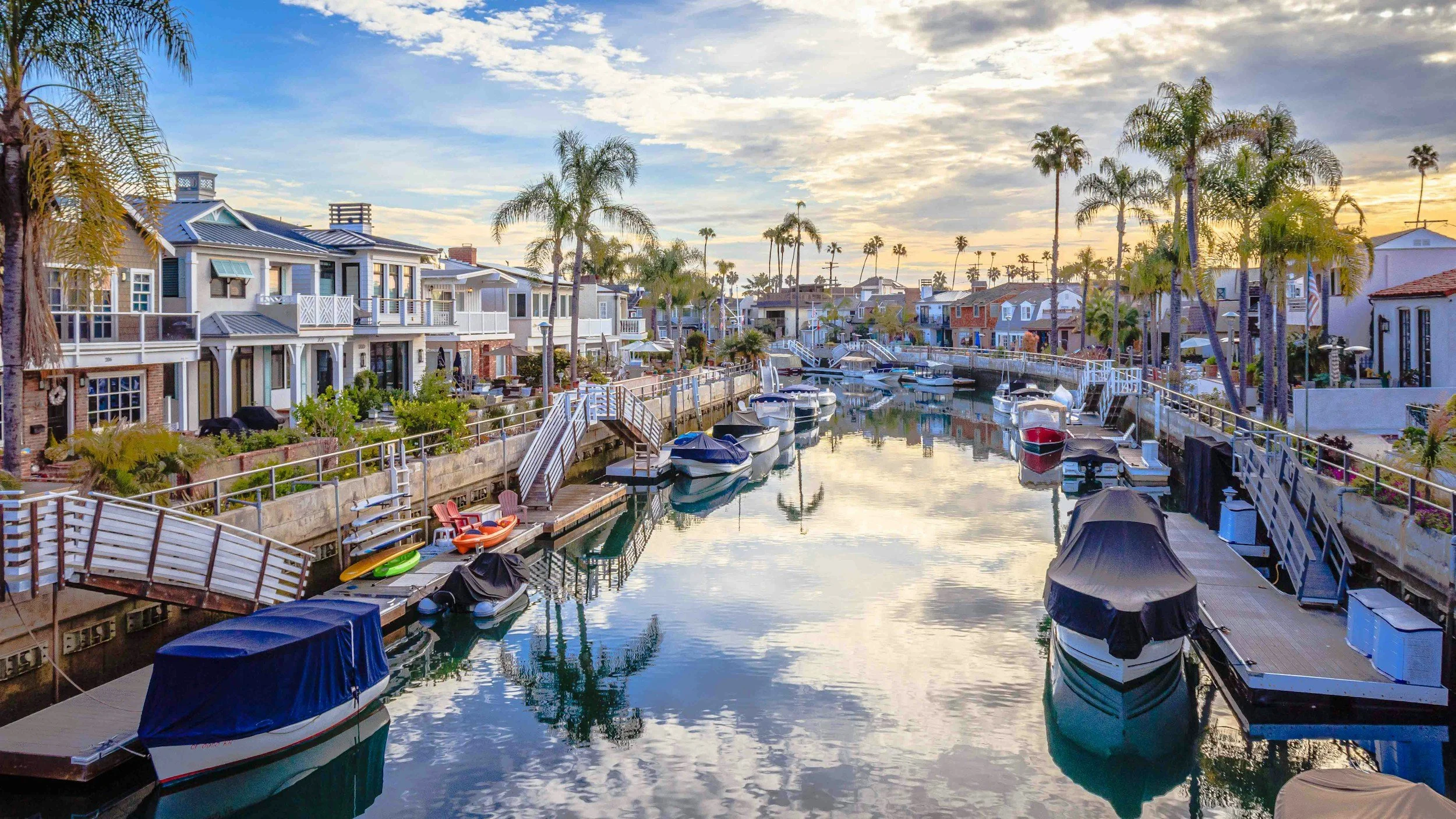 Residential houses along a canal with boats docked, palm trees, and a sunset sky.