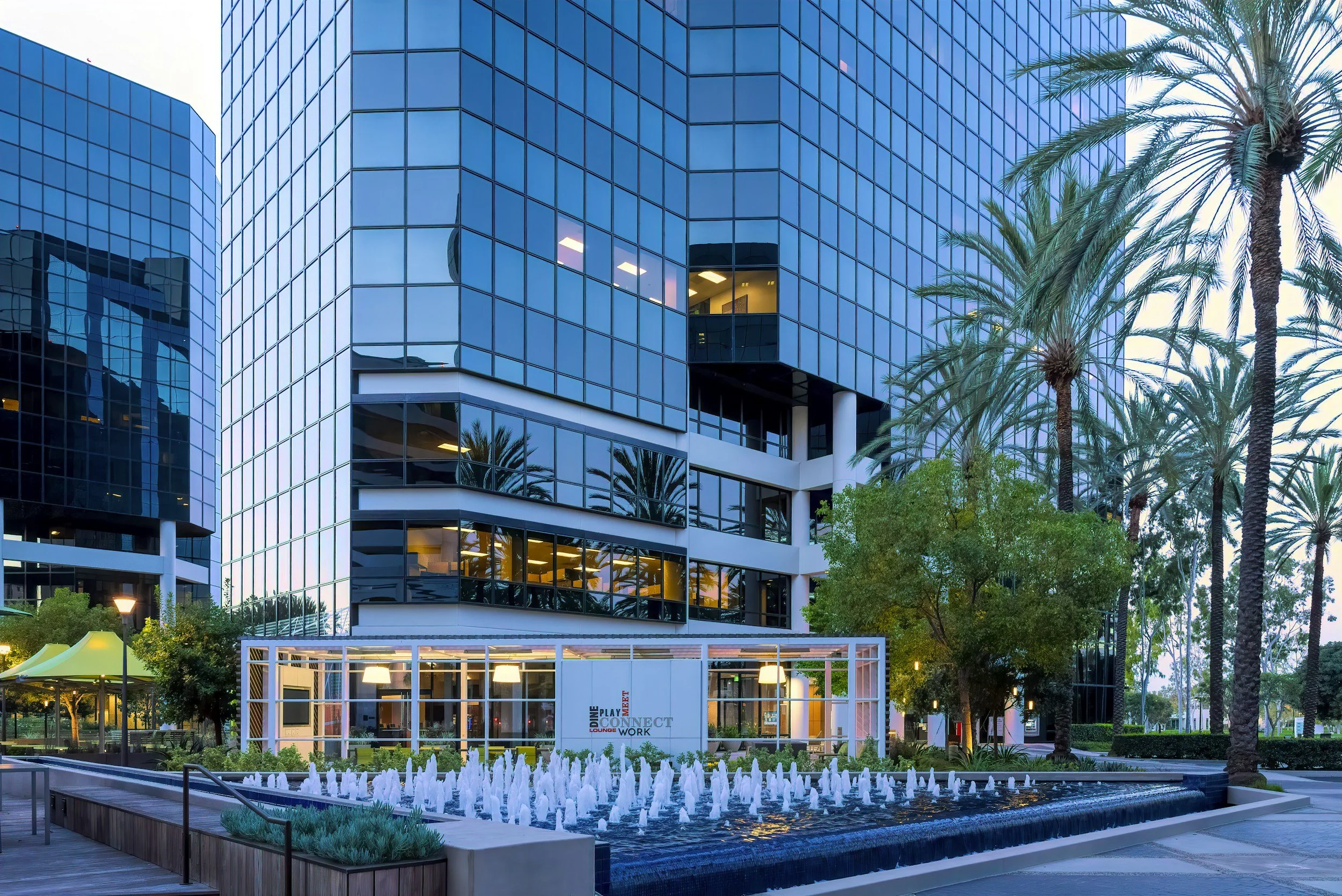 Modern glass office building with reflected windows, palm trees, and a water fountain in the foreground.