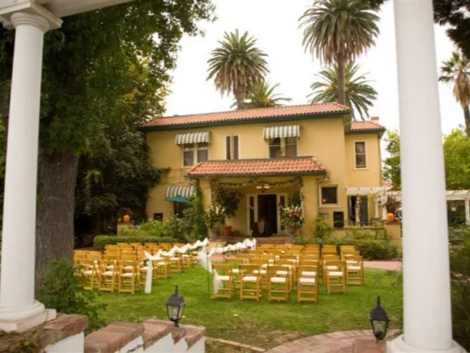 The French Estate in Orange, a yellow two-story house with red tile roof, striped awnings, set in a lush garden with tall palms, chairs arranged for an outdoor event, and white fabric decorations.