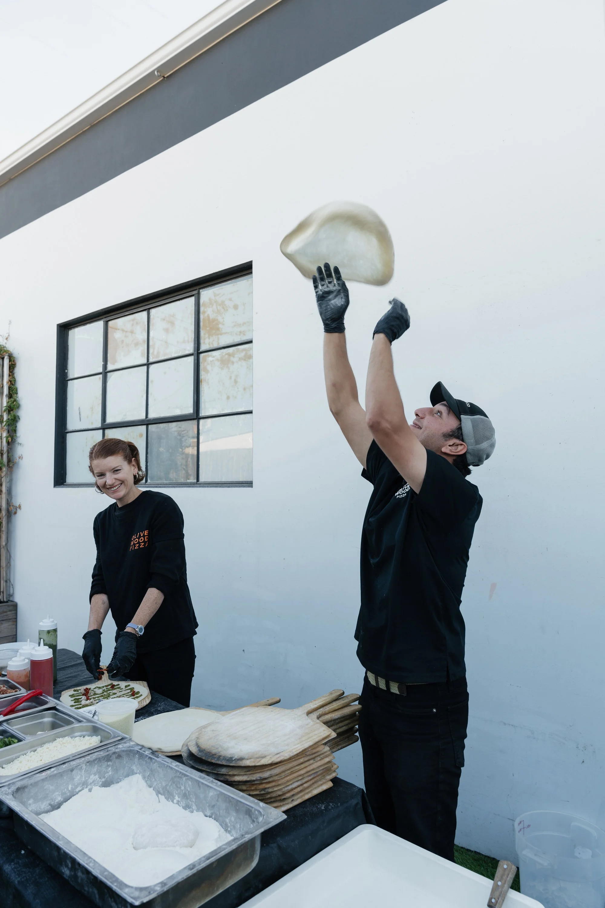 Two people, a man and a woman, preparing pizza outdoors in front of a white wall. The man is tossing dough in the air while the woman is preparing ingredients on a table. The table has various pizza toppings and baking supplies.