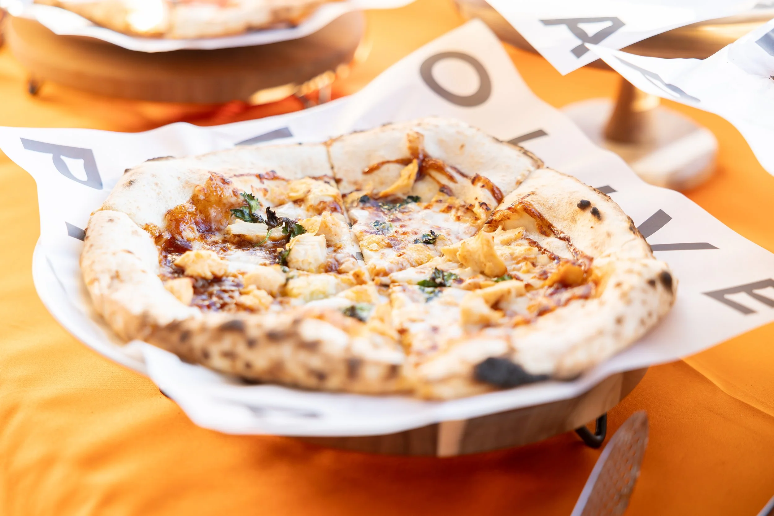 A Barbeque Chicken pizza with mozzarella cheese, chicken, and cilantro on white wax paper with "OLIVE WOOD PIZZA" printed in black ink, atop a white china plate on a wooden riser, on an orange table cloth.
