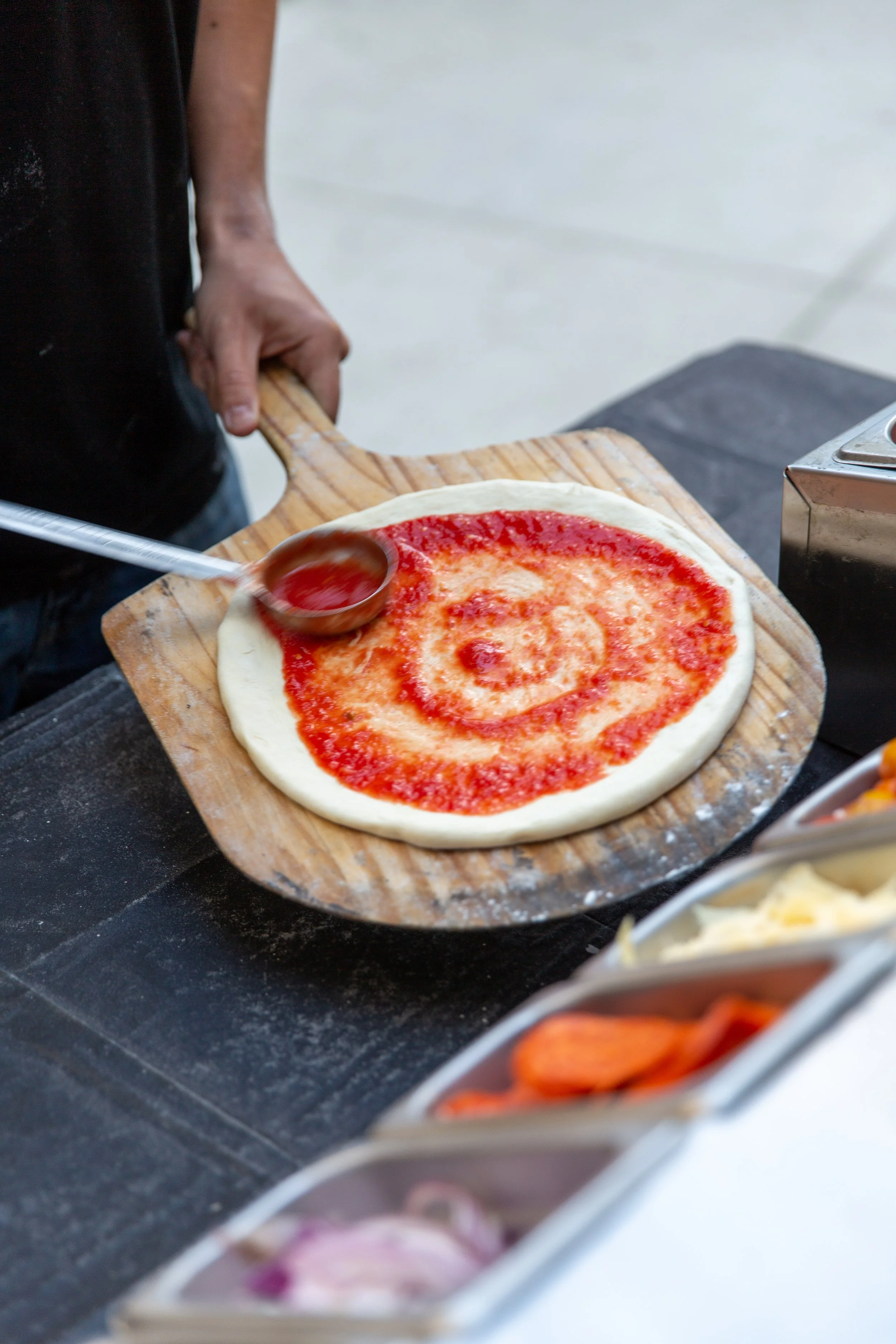 A person spreading crushed tomato on raw pizza dough on a wooden pizza peel.