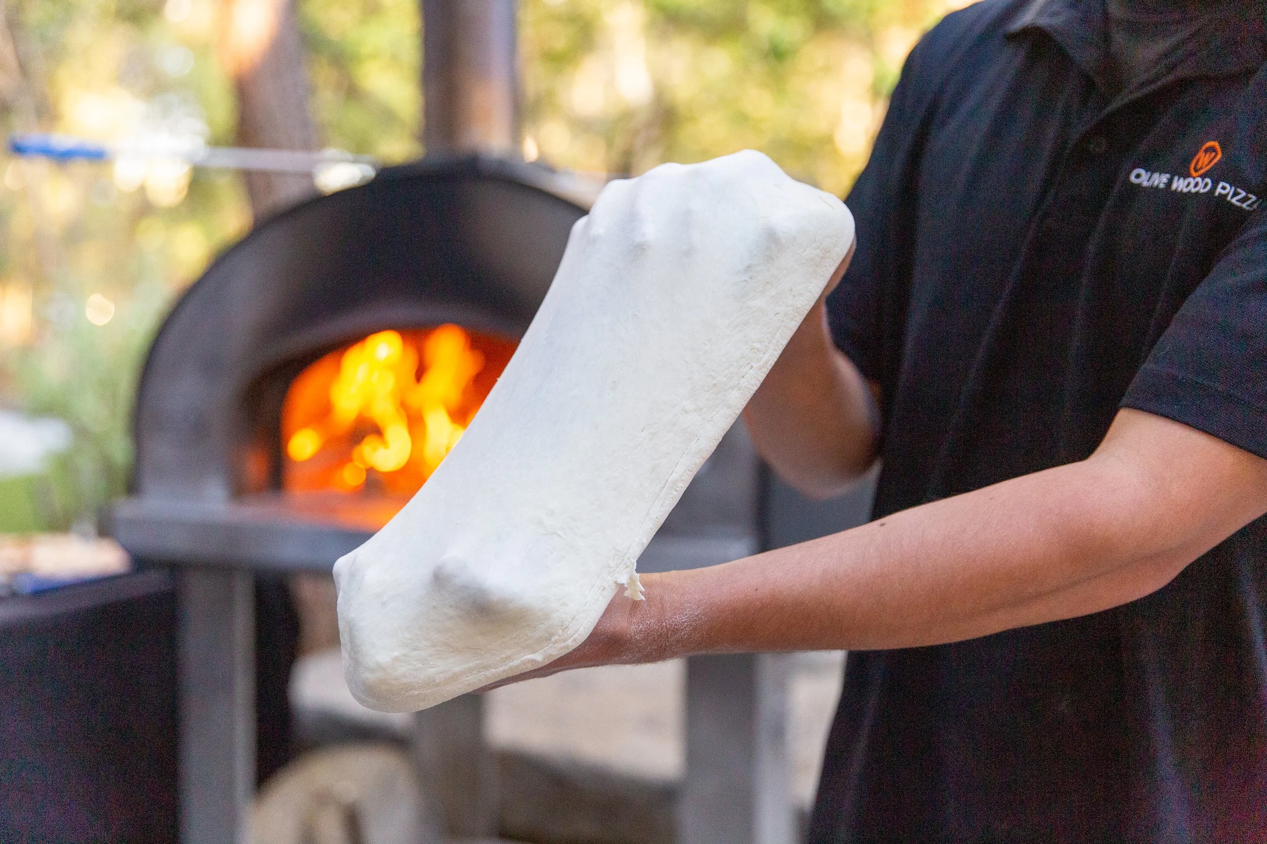 Fresh pizza dough being hand-stretched in front of a wood-fired oven by Olive Wood Pizza